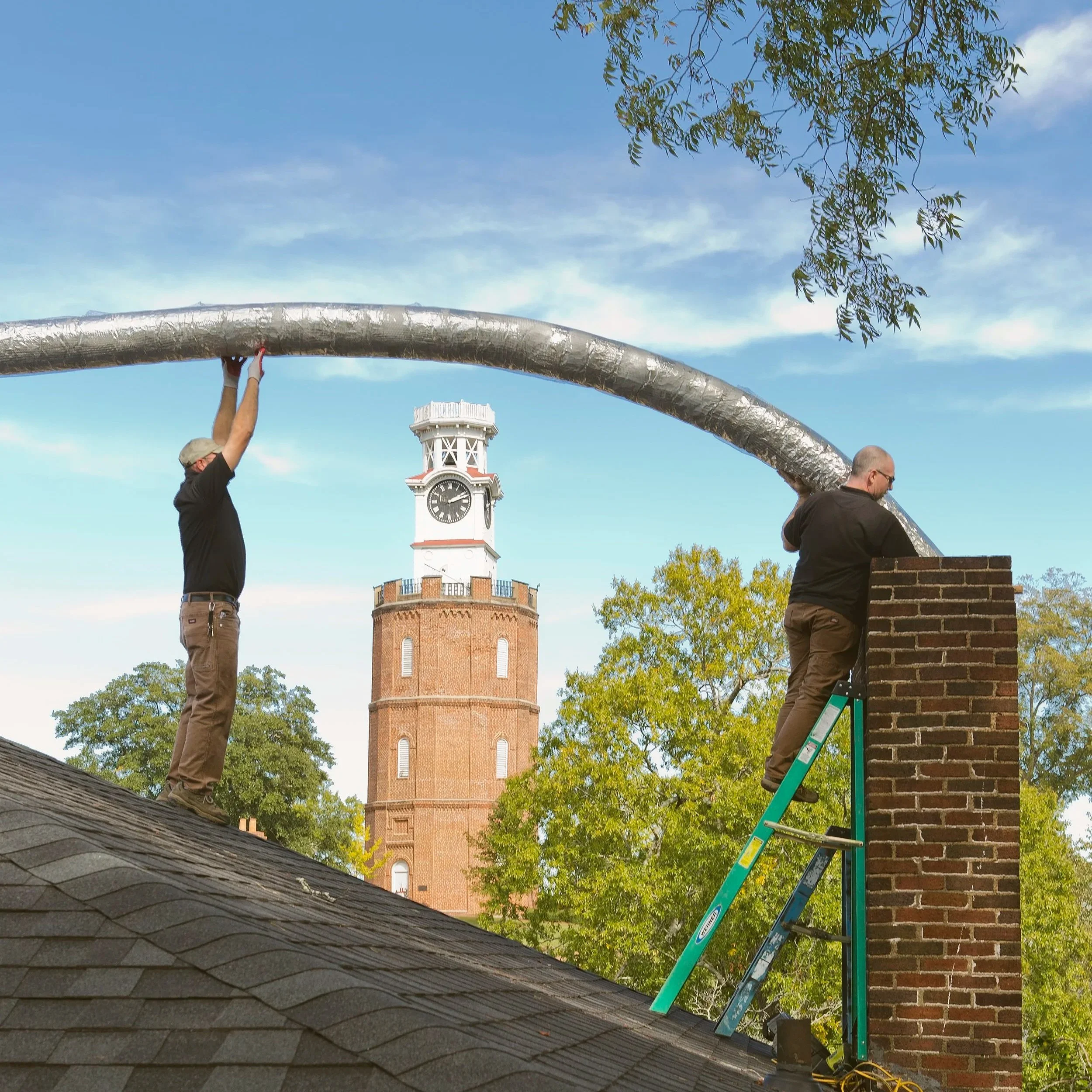 Two men working on chimneys on a rooftop, with a clock tower and trees in the background under a blue sky.
