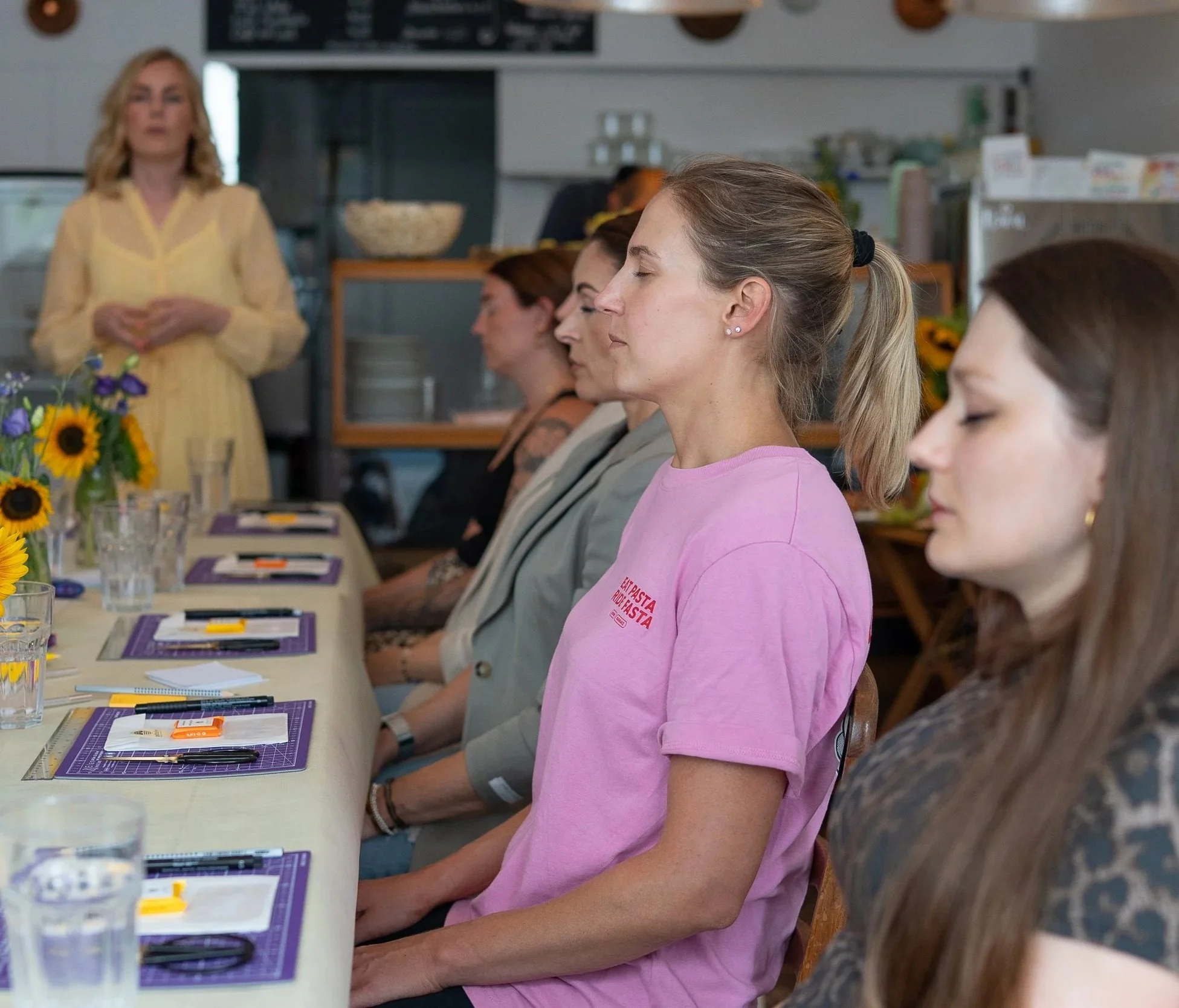 Menschen sitzen nebeneinander am Tisch und haben die Augen geschlossen, so als ob sie gerade an einer Meditation teilnehmen. Im Hintergrund ist die Workshopleiterin im gelben Kleid zu sehen.