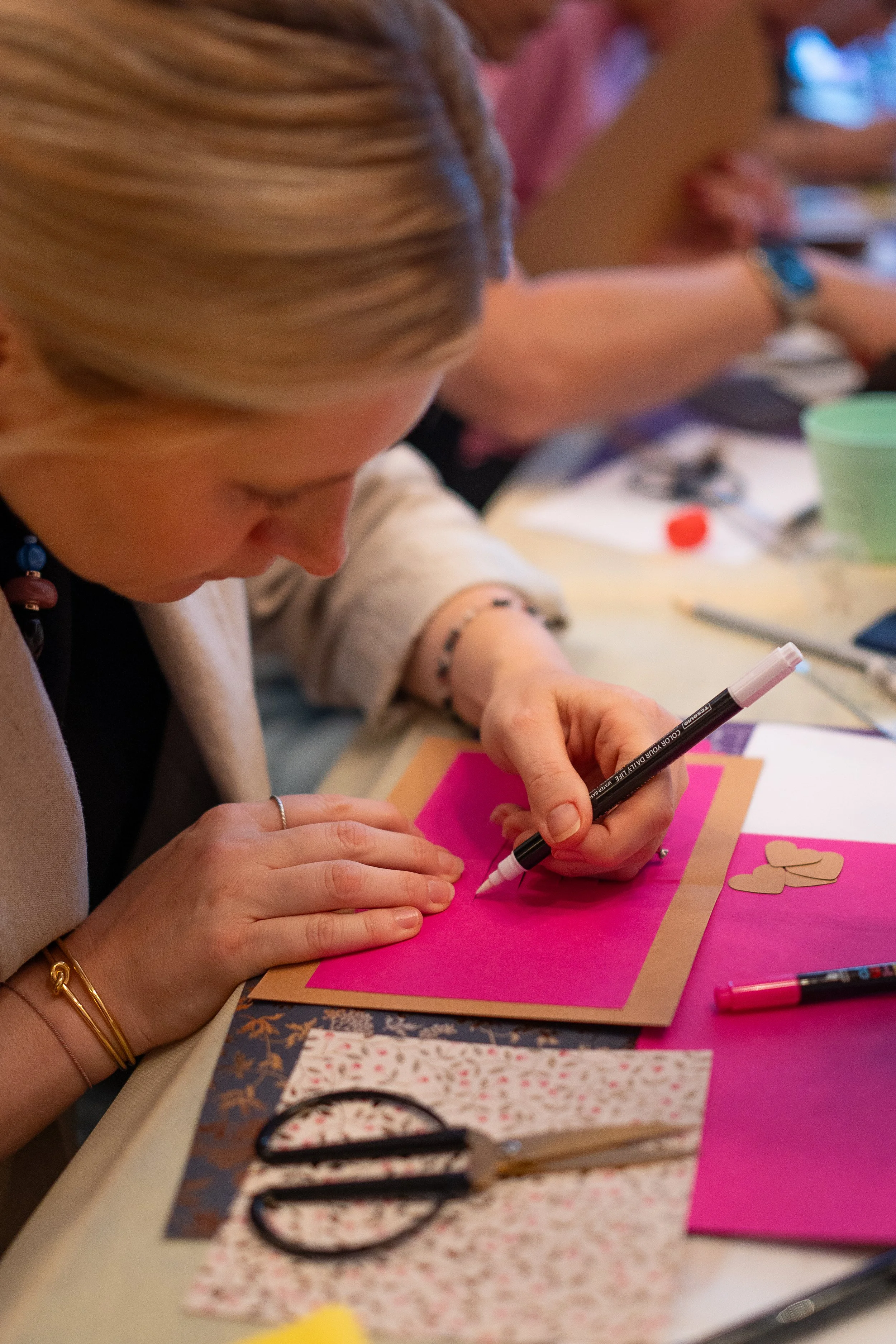 Person erstellt eine Grußkarte mit buntem Papier, Schneiderschere und Stiften auf einem Tisch. Sie malt gerade etwas mit einem weißen Stift auf pinken Papier.