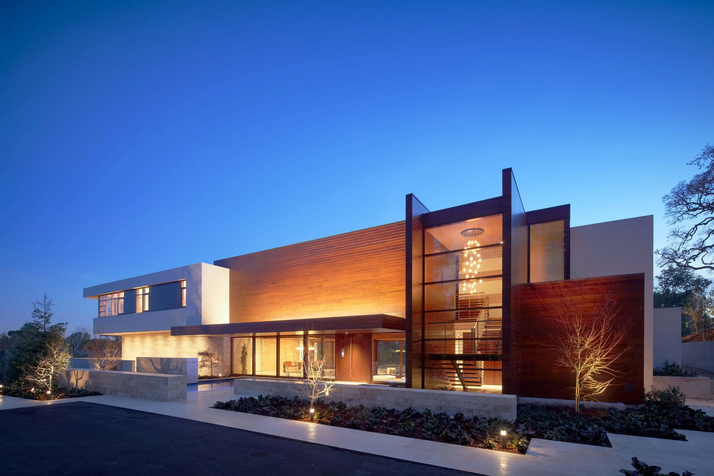 Silicon Valley residence exterior at night, illuminated wood facade and glass stairwell with pendant chandelier