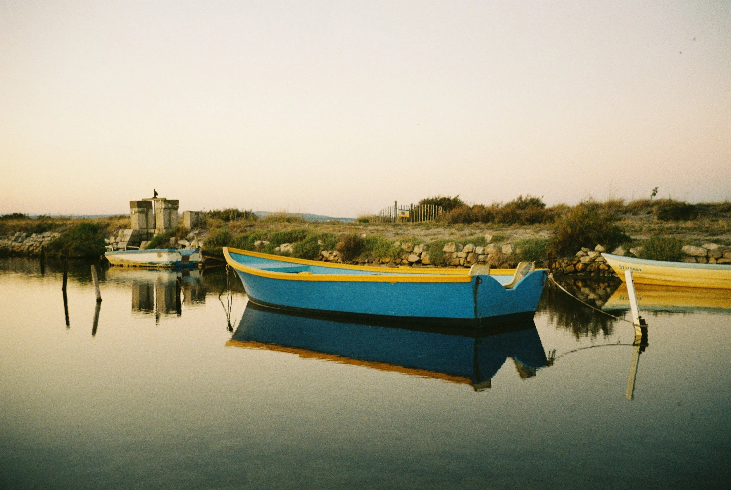 A blue and orange wooden boat reflected in calm water, near a stone-lined shore with some vegetation and small structures under a clear sky.