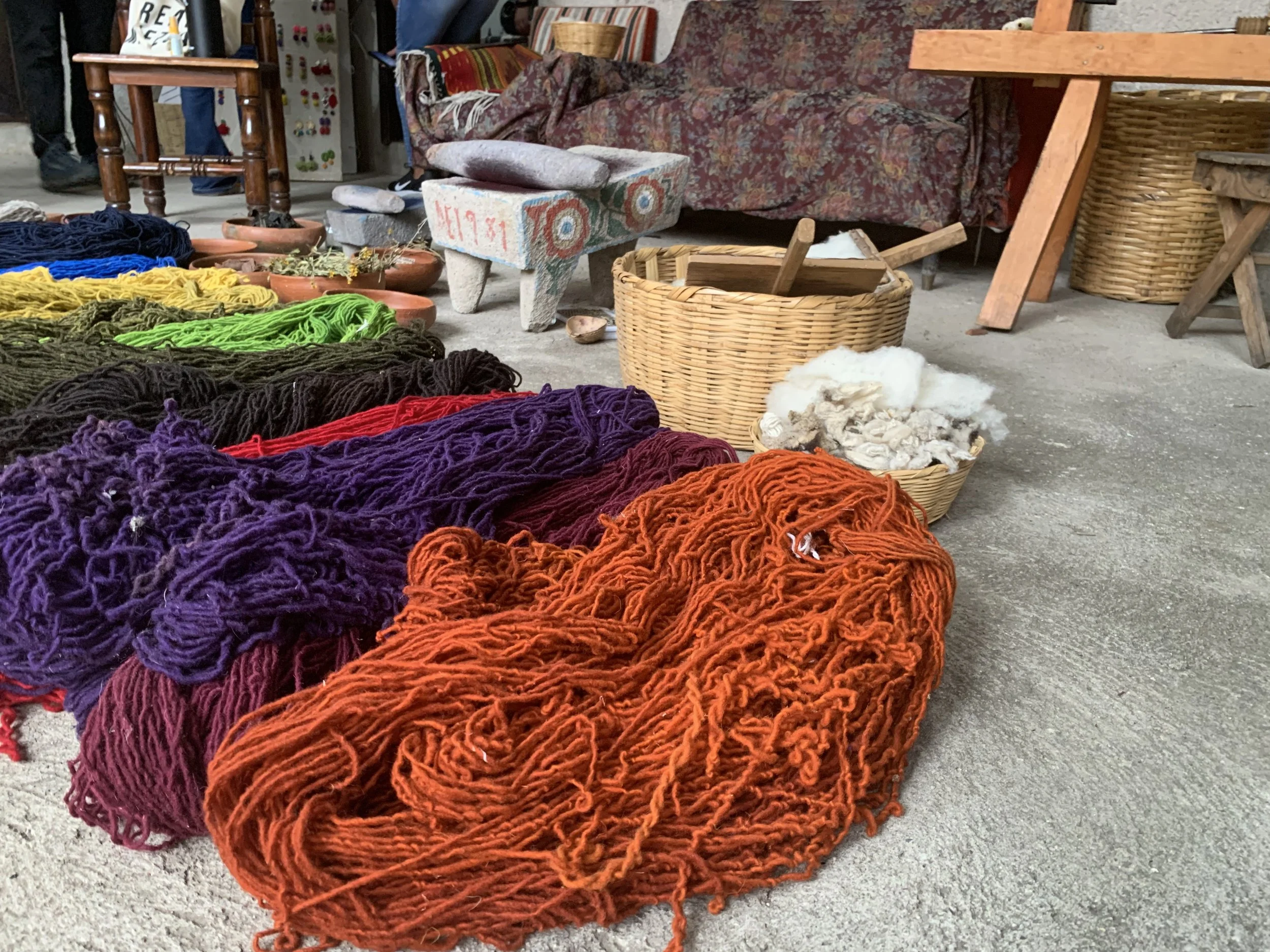Colorful skeins of yarn on the ground, including orange, purple, and red, near baskets and rustic furniture in a workshop setting.