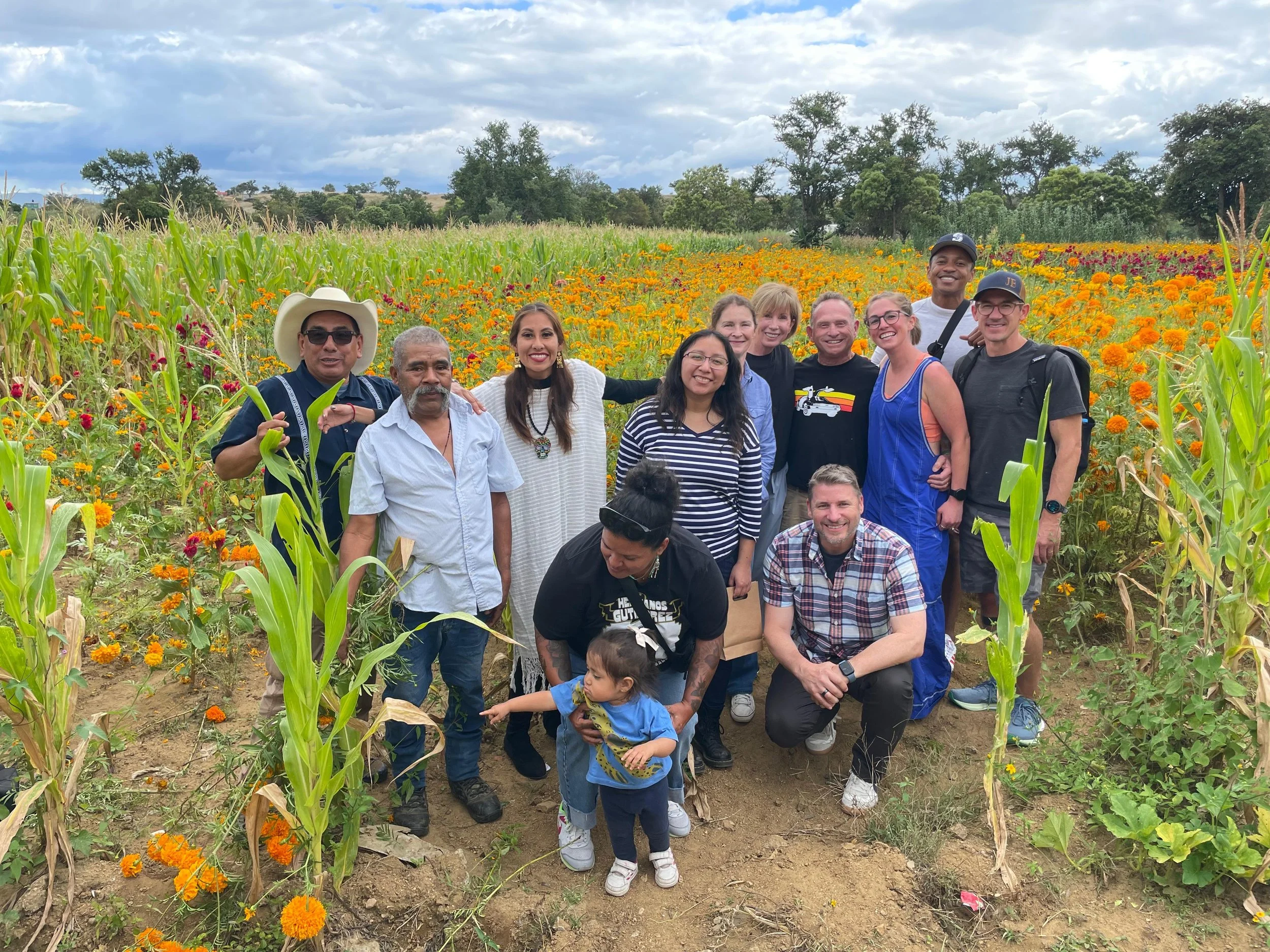 Group of people posing in a field of marigold flowers and corn plants, with a cloudy sky in the background.