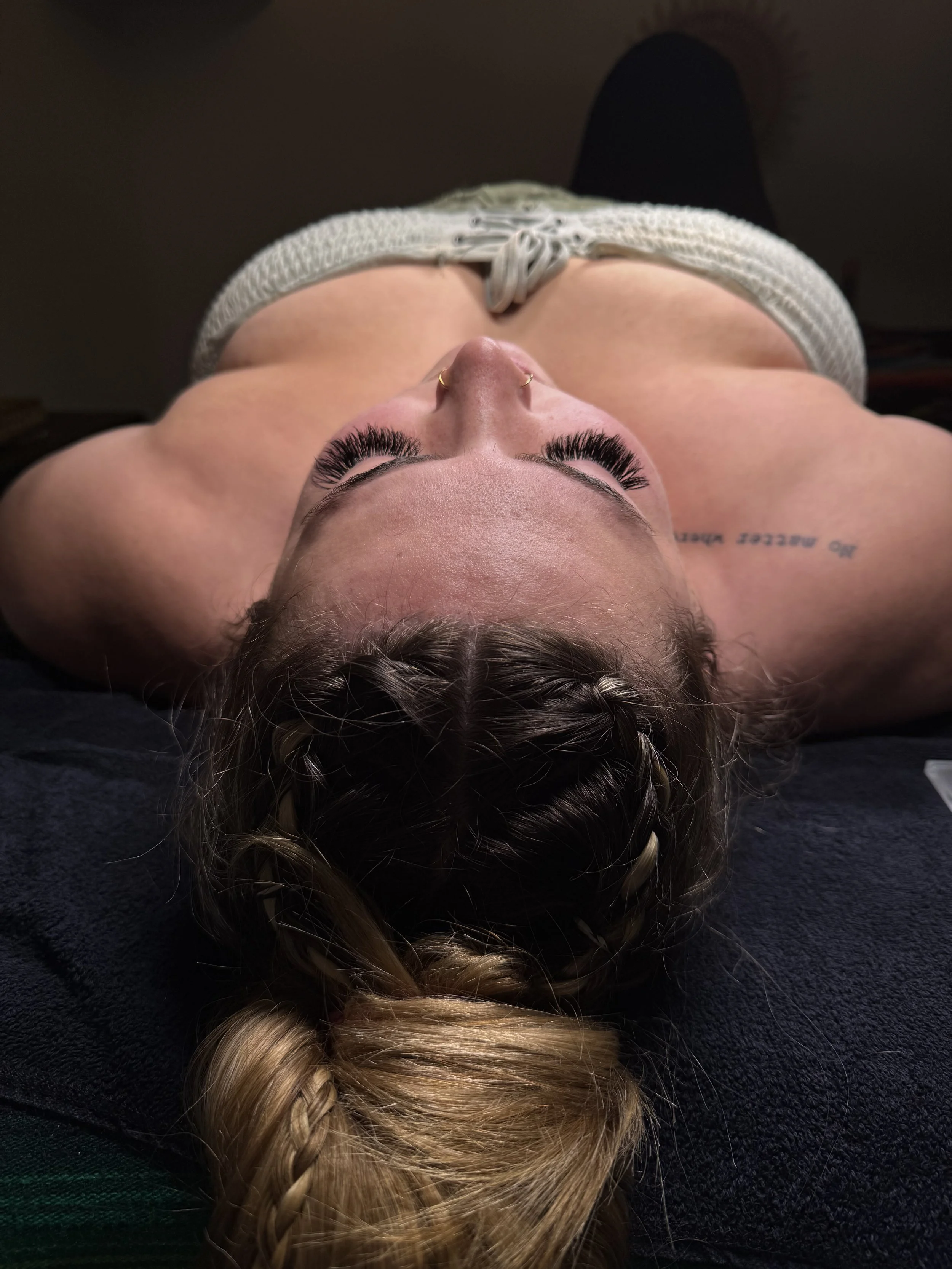 A woman is lying on her back, looking up, with long blonde hair styled in a braid. She has dark eye makeup, including thick eyelashes, and is wearing a septum piercing. She is dressed in a light-colored knitted top with a drawstring at the front.