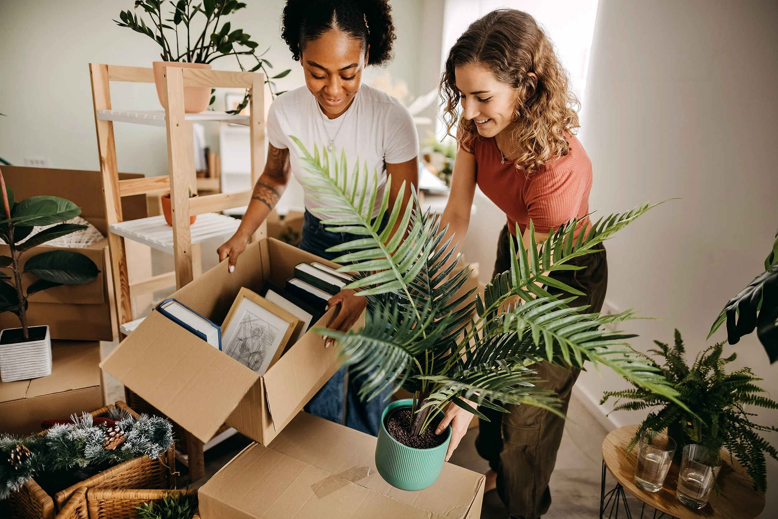 Two women are unpacking a box of framed photos and books, surrounded by plants in a well-lit room.