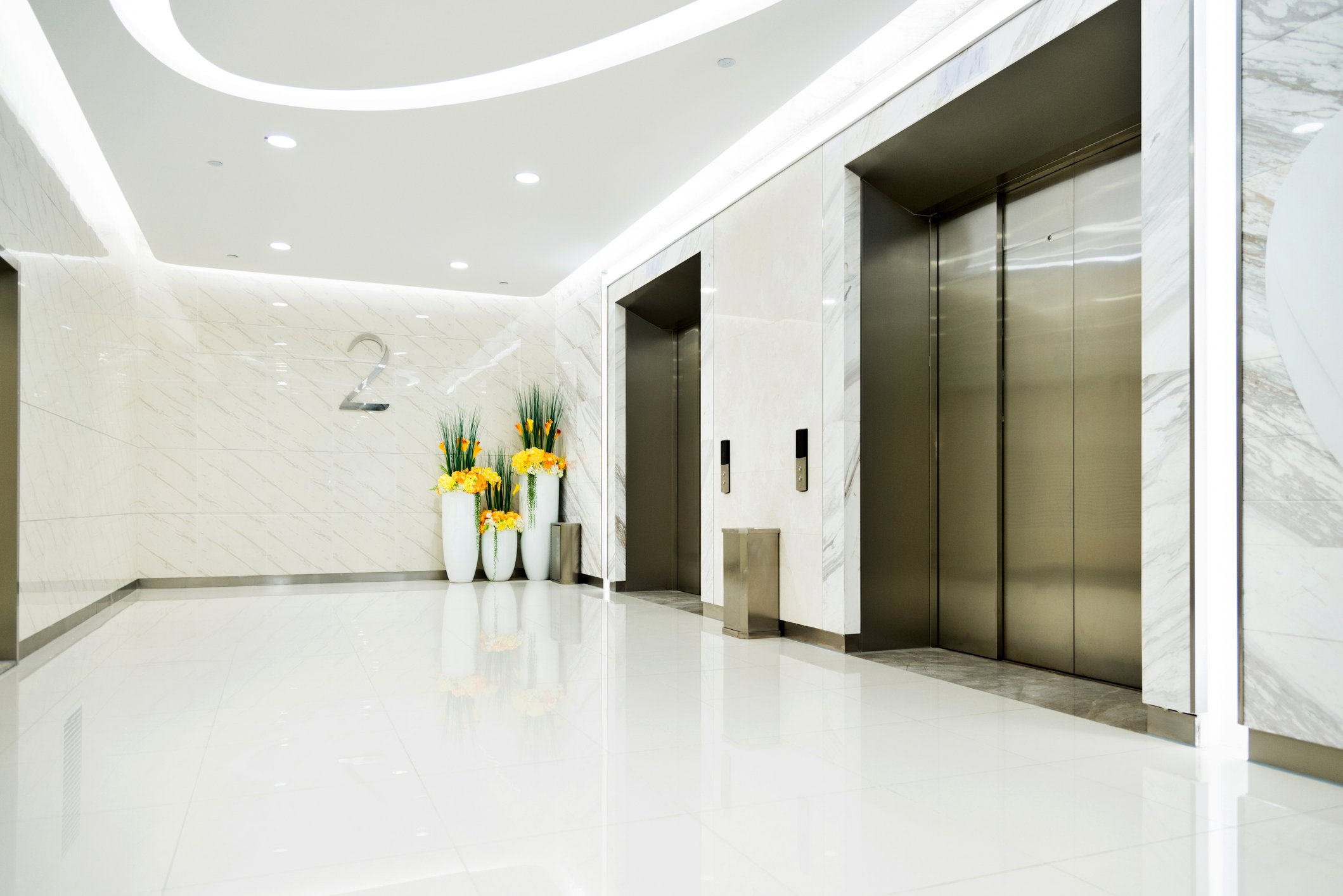 Modern hotel lobby with two elevators, white marble walls and floors, and decorative yellow and orange flowers in tall white vases.