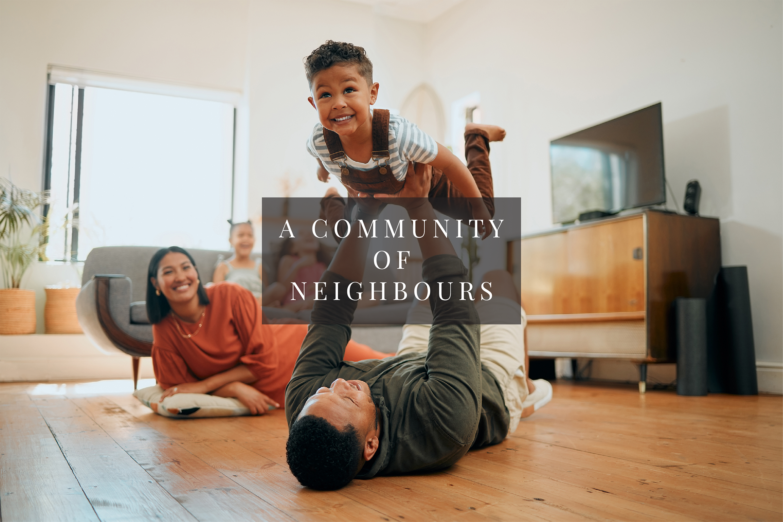 A family playing on the living room floor, with a boy being lifted by a man, and a woman and girl smiling in the background.