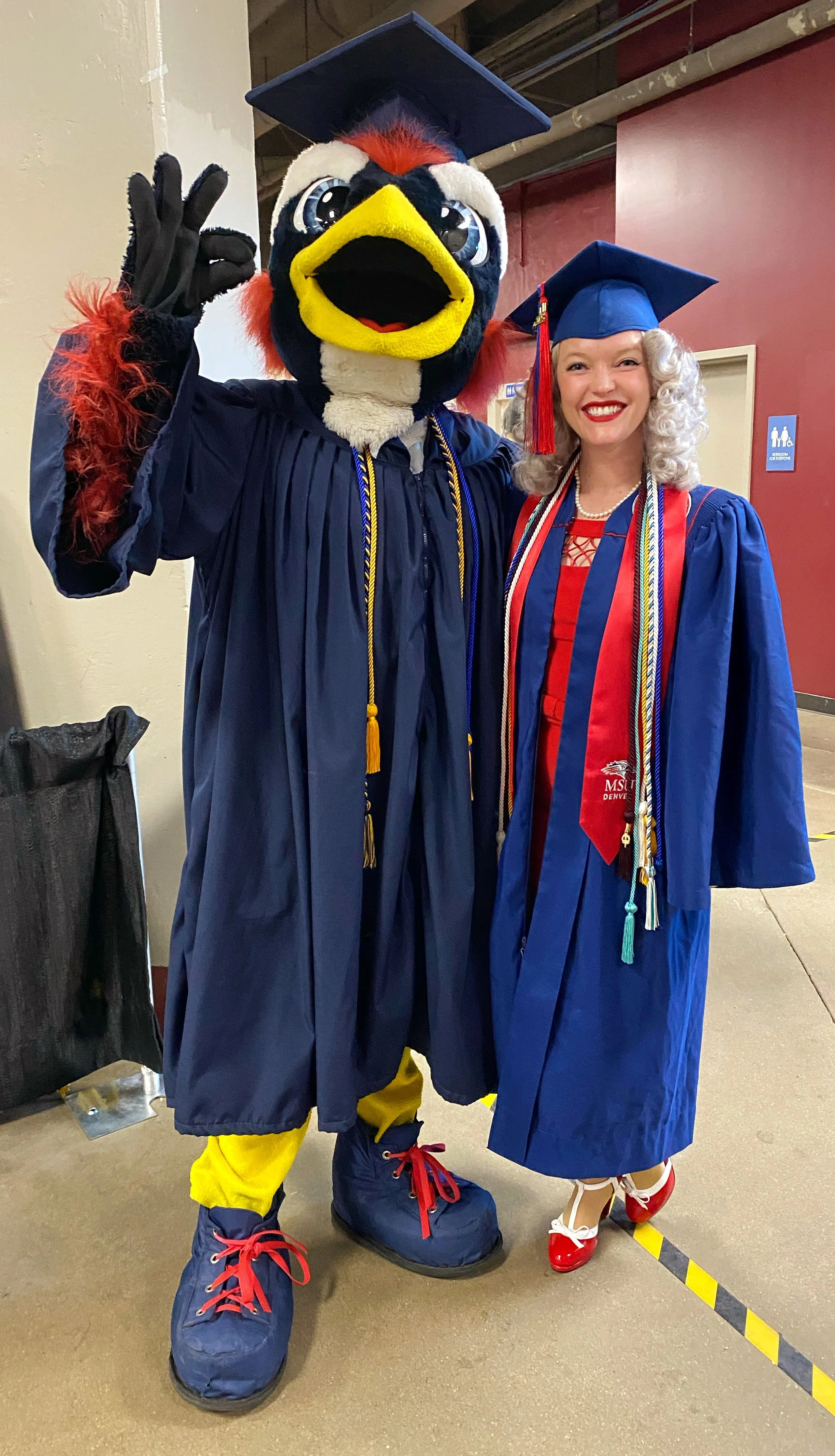 Whitney Haddad at her MSU Denver graduation, wearing a cap and gown with honor cords, standing next to the university's mascot.