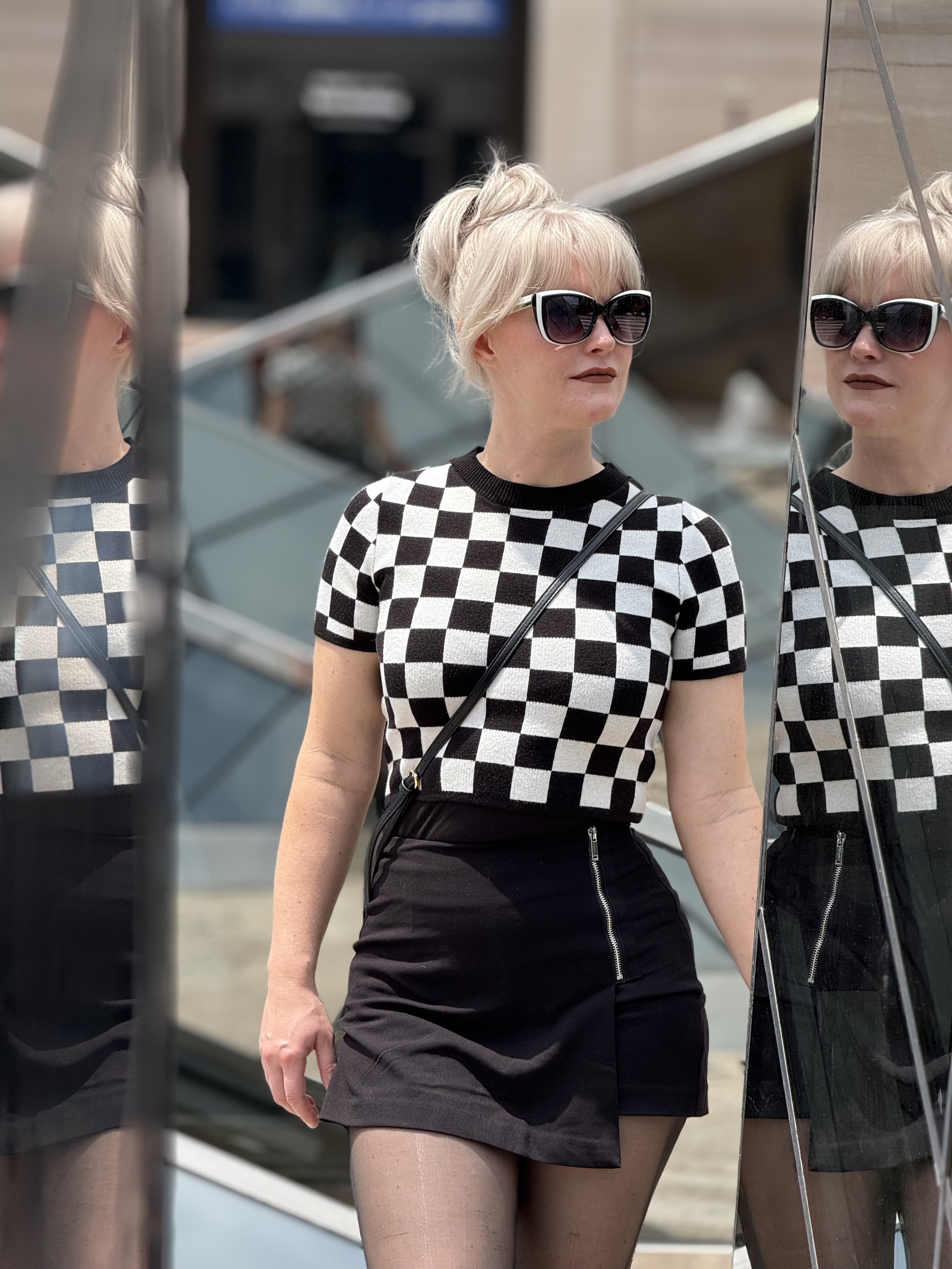 Whitney Haddad standing outdoors near a mirrored glass structure, reflections visible on either side.