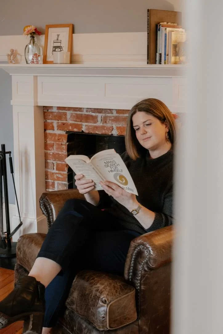 A woman sitting on a brown leather chair reading a book in front of a brick fireplace, with decorations and books on the mantel.