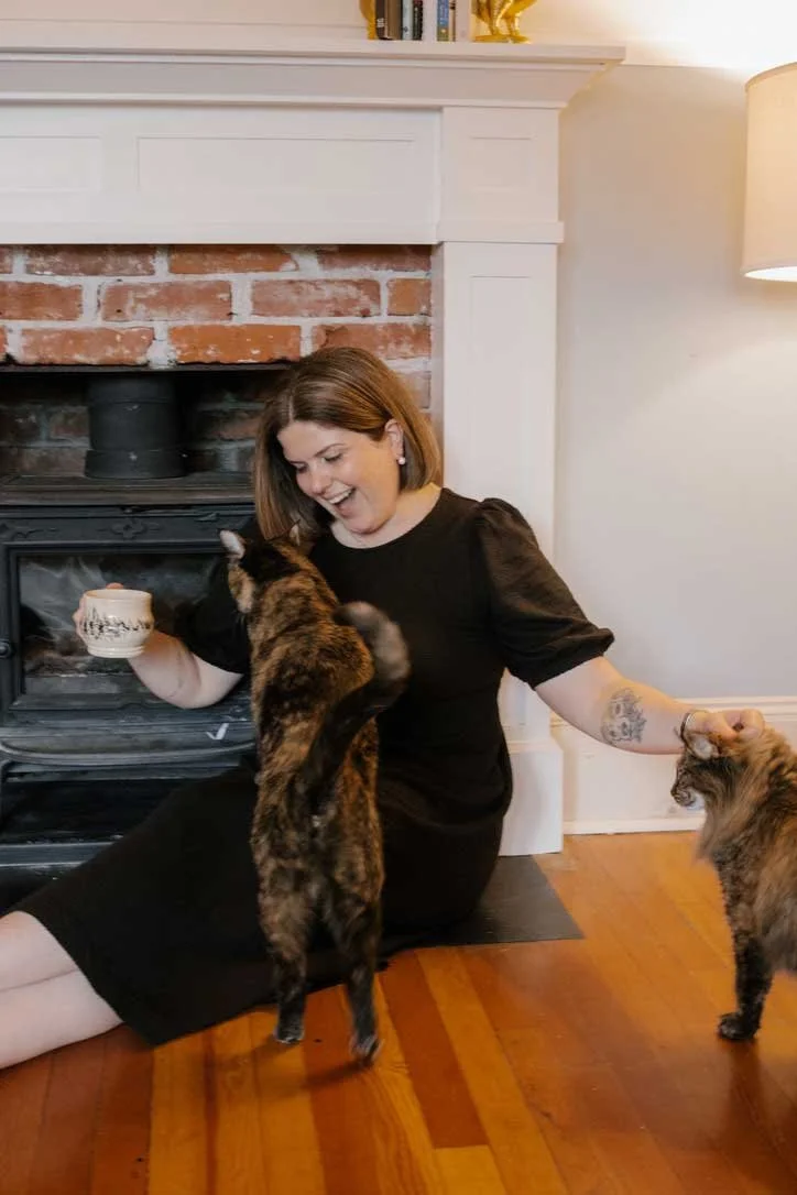 Woman sitting on the floor near a fireplace, holding a mug, with two cats, one standing and one being petted.