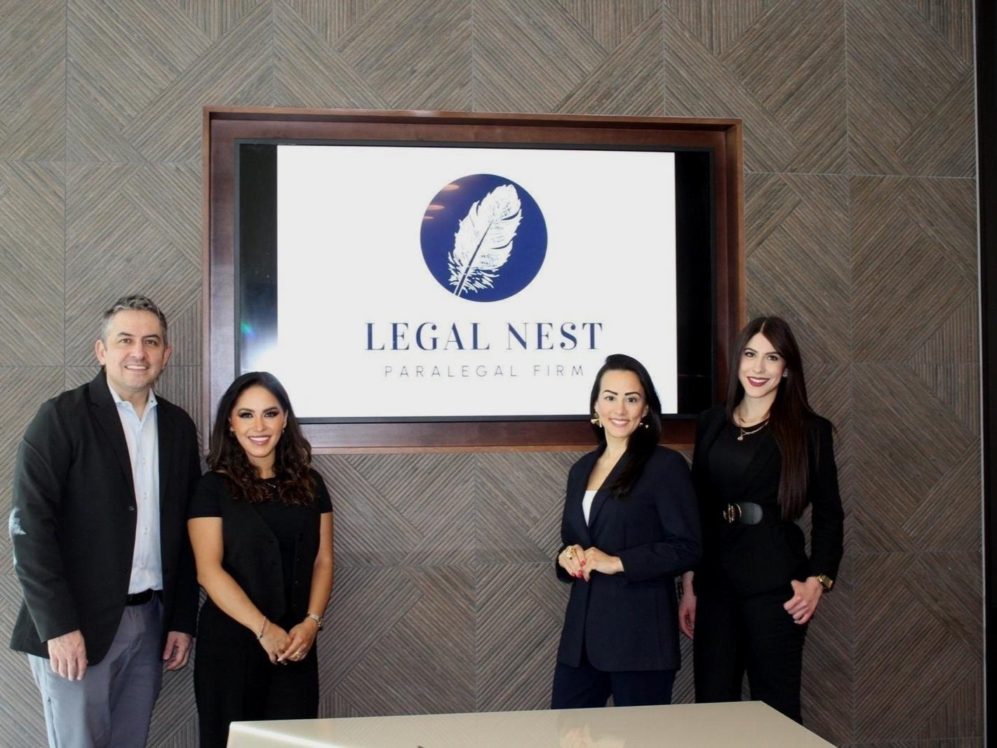 Un grupo de cuatro personas, tres mujeres y un hombre, posan frente a un letrero que dice "Legal Nest Paralegal Firm" con un logo de pluma. Todos visten ropa profesional y están sonrientes. La pared de fondo tiene un diseño geométrico.