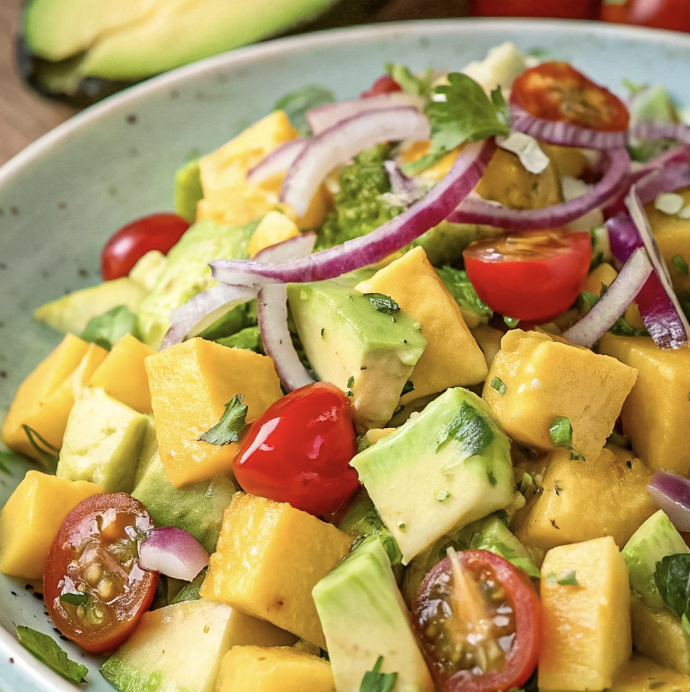 Close-up of a fresh salad with avocado, cherry tomatoes, mango chunks, red onions, and green herbs in a blue bowl.