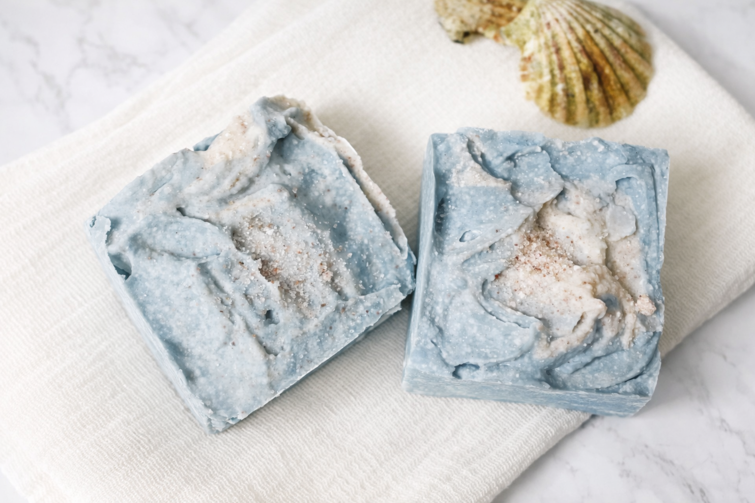 Two light blue and white marbled soap bars with salt and exfoliants, placed on a white textured cloth with a seashell nearby.