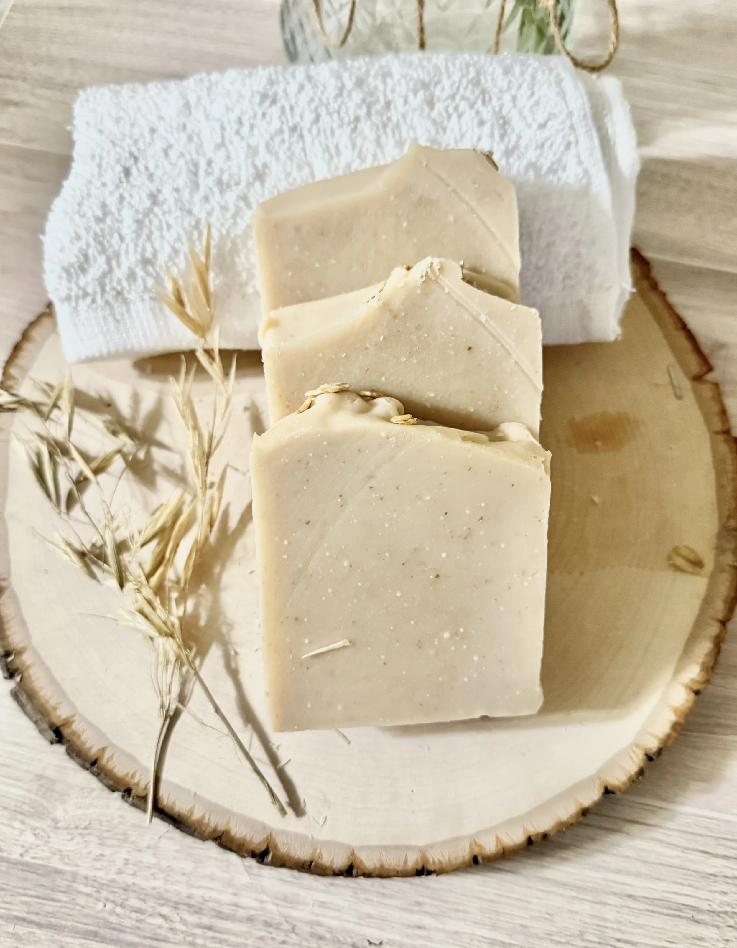 Three bars of beige handmade soap on a wooden tray with a dried wheat stalk, a white towel, and some greenery in the background.