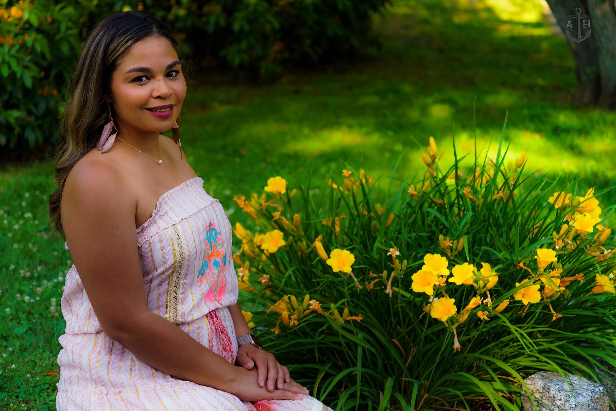 A woman sitting outdoors near a bush of yellow flowers, smiling at the camera.