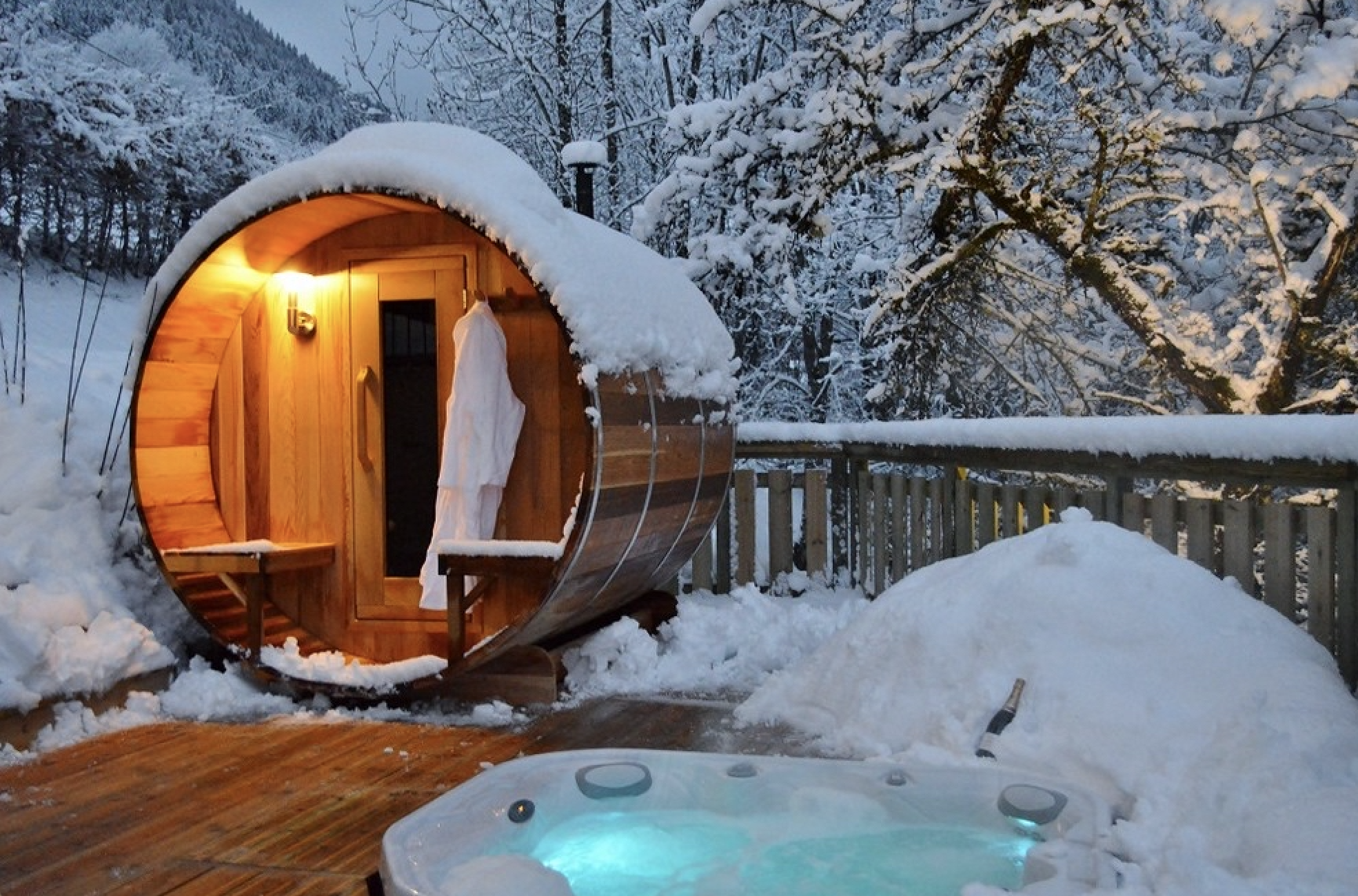 Snow-covered outdoor scenery with a small wooden sauna, warm interior light, and a hot tub with bubbling water in snowy surroundings and trees.