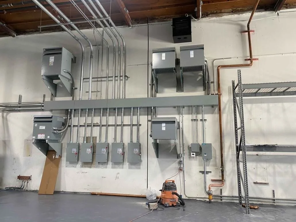 Electrical wiring, conduit pipes, control panels, and boxes mounted on a warehouse wall with a vacuum cleaner on the floor.