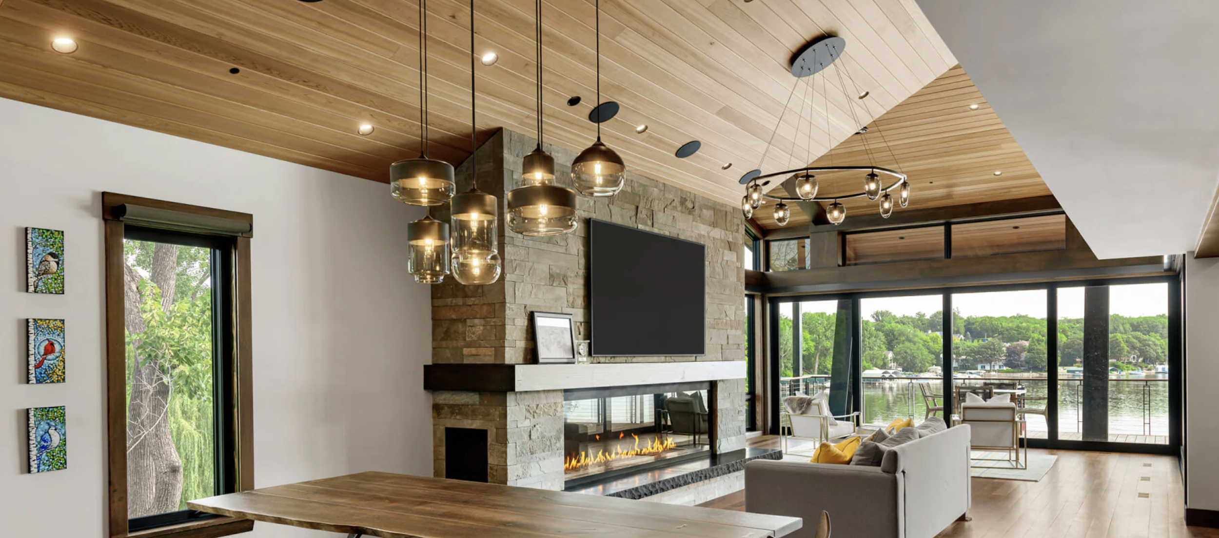 Modern living room with large glass doors overlooking water, featuring a stone fireplace with a mounted TV, wooden ceiling with pendant lights, and seating area.