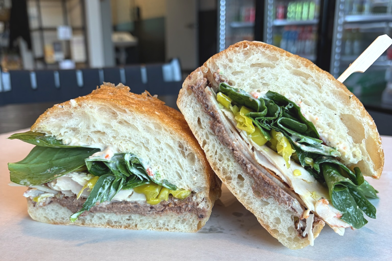 Close-up of a sandwich with leafy greens, cheese, and sliced meat on a toasted bun, on a paper-lined surface in a cafe setting