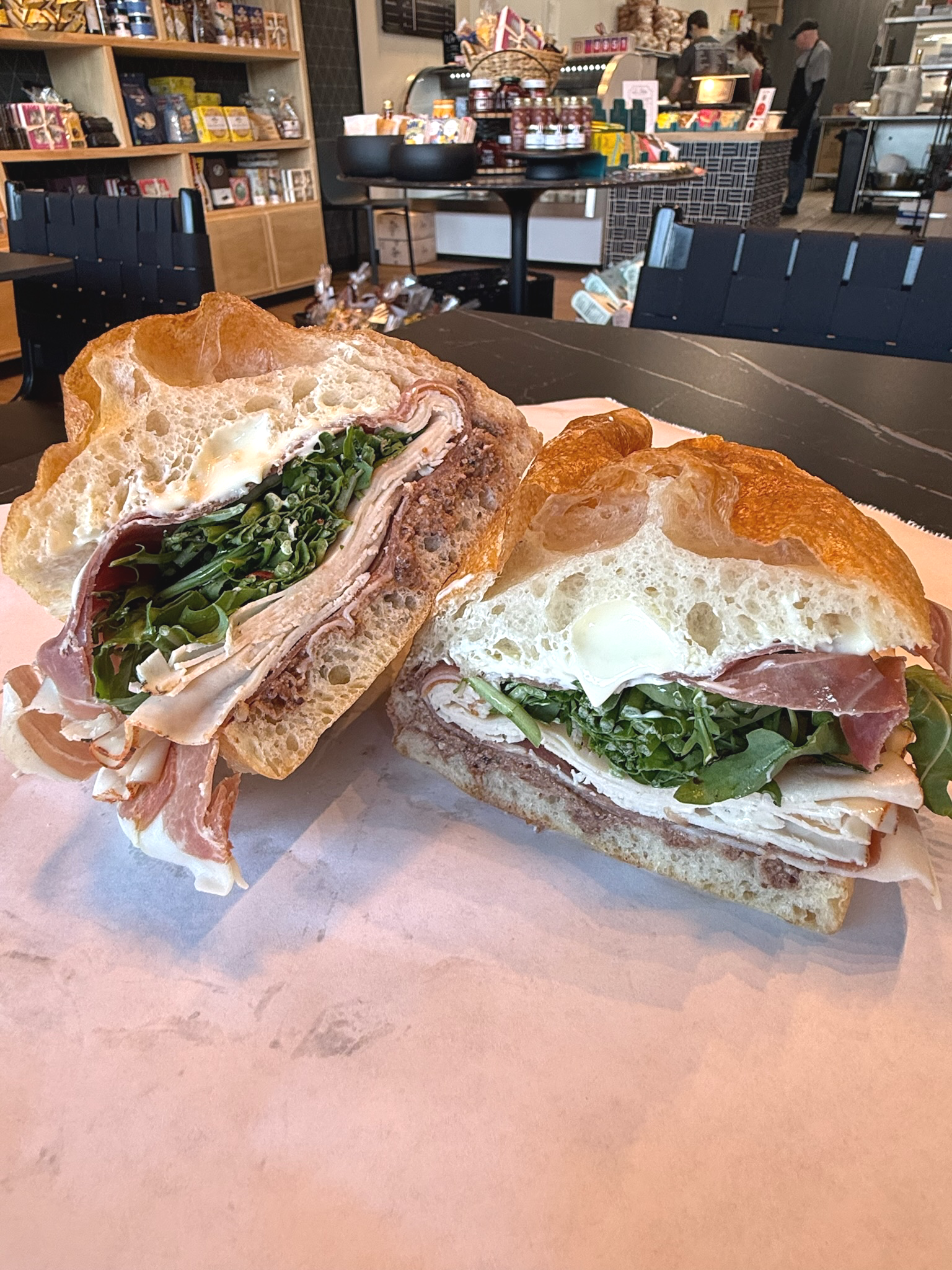 A sandwich with layers of turkey, ham, turkey, cheese, arugula, and a fried egg on a baguette, served on pink paper, in a café or bakery with shelves of products and customers in the background.