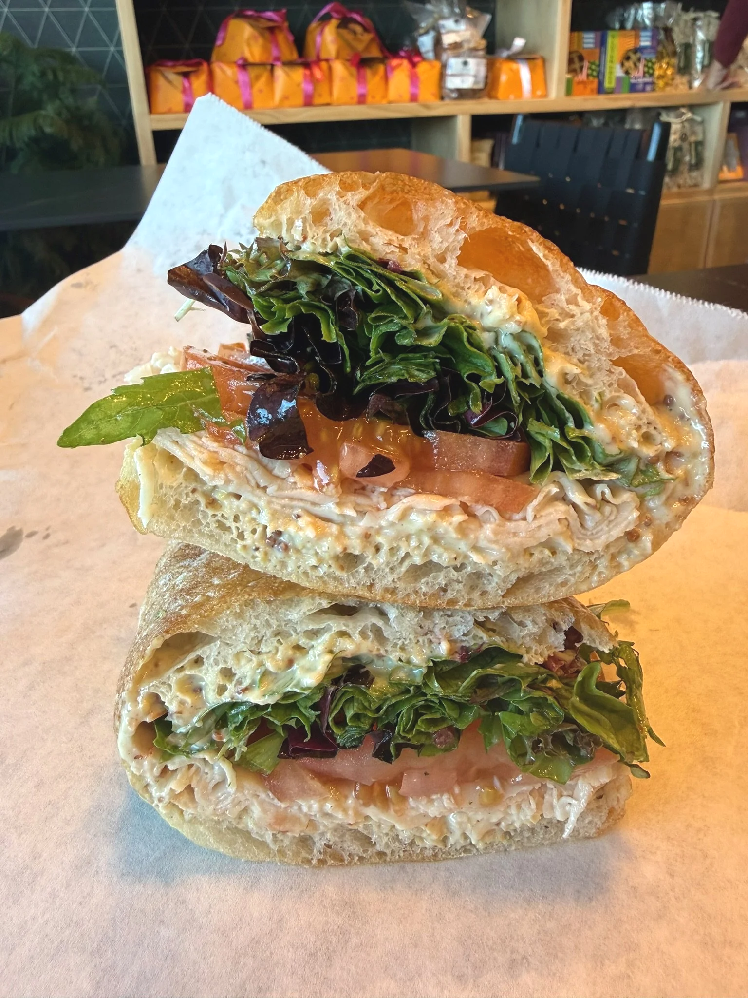 Close-up of a sandwich with lettuce, tomato, and creamy filling, cut in half and placed on parchment paper in a cafe or sandwich shop setting.