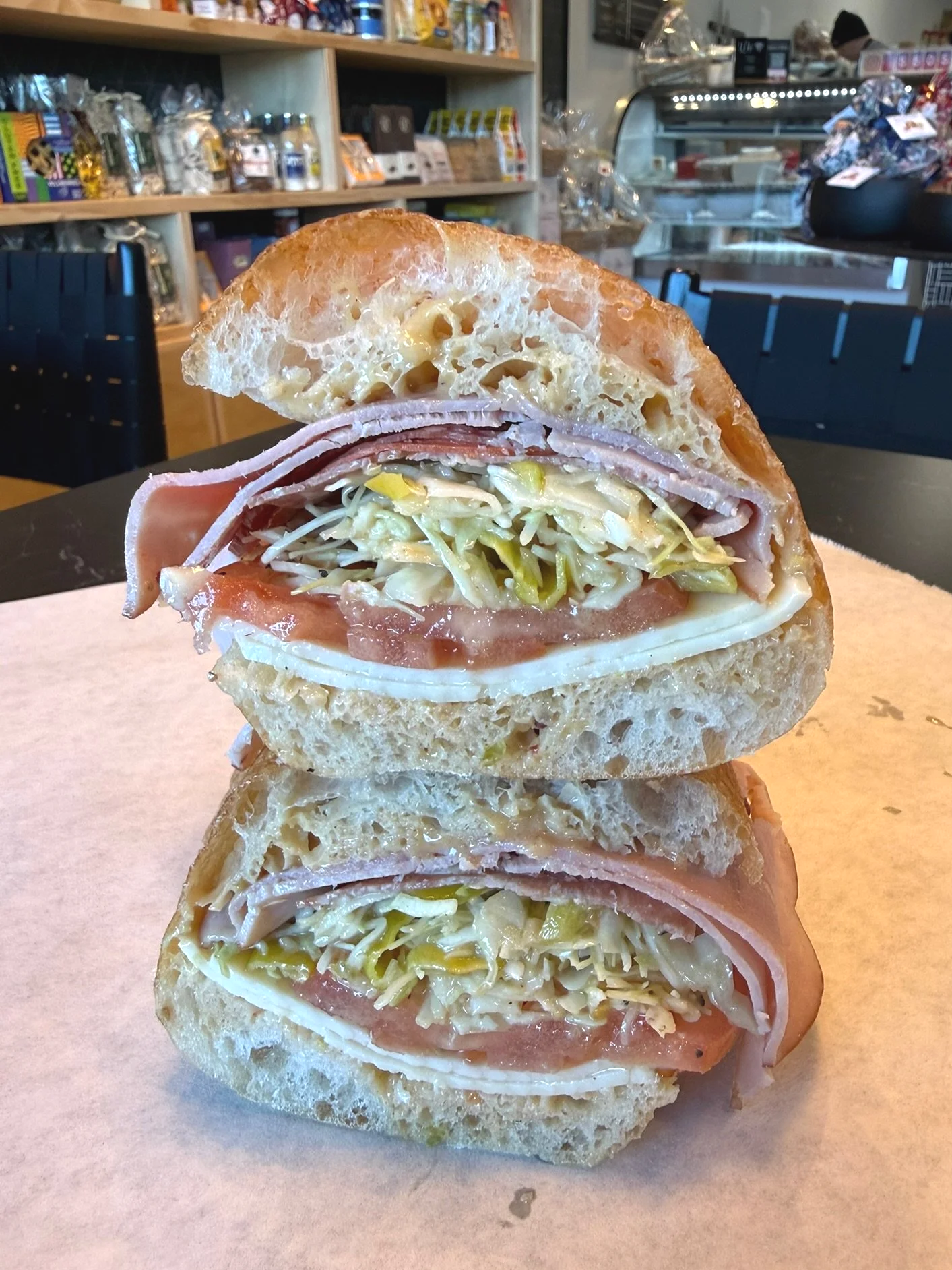 Sliced sandwich with bread, deli meat, shredded lettuce, tomato, cheese, and other fillings, placed on a piece of parchment paper in a cafe setting