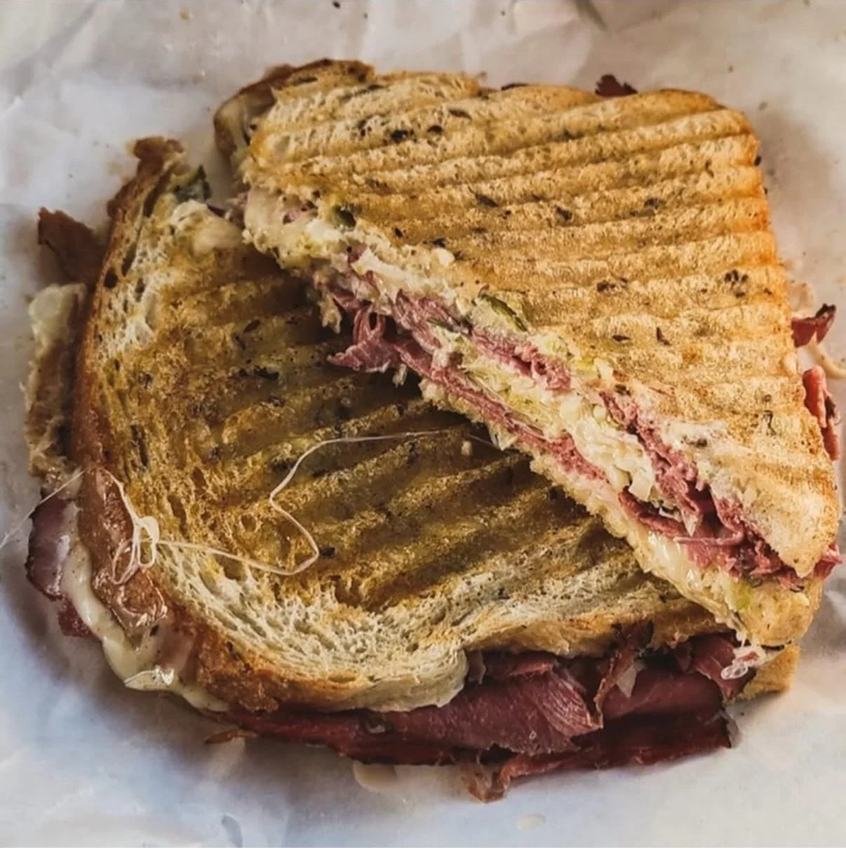 Close-up of a grilled sandwich with meat and melted cheese, cut in half to show ingredients inside, on parchment paper.