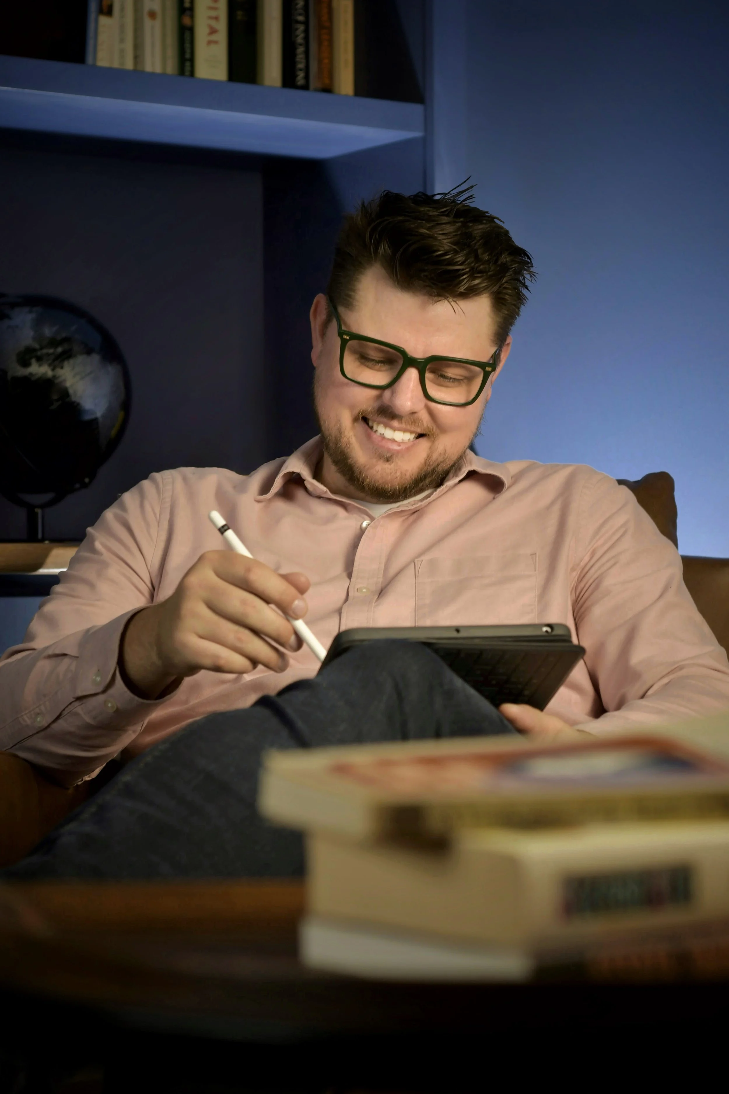 A man with glasses is sitting on a couch, smiling, while using a stylus on a tablet. There are books and a globe on a shelf behind him.