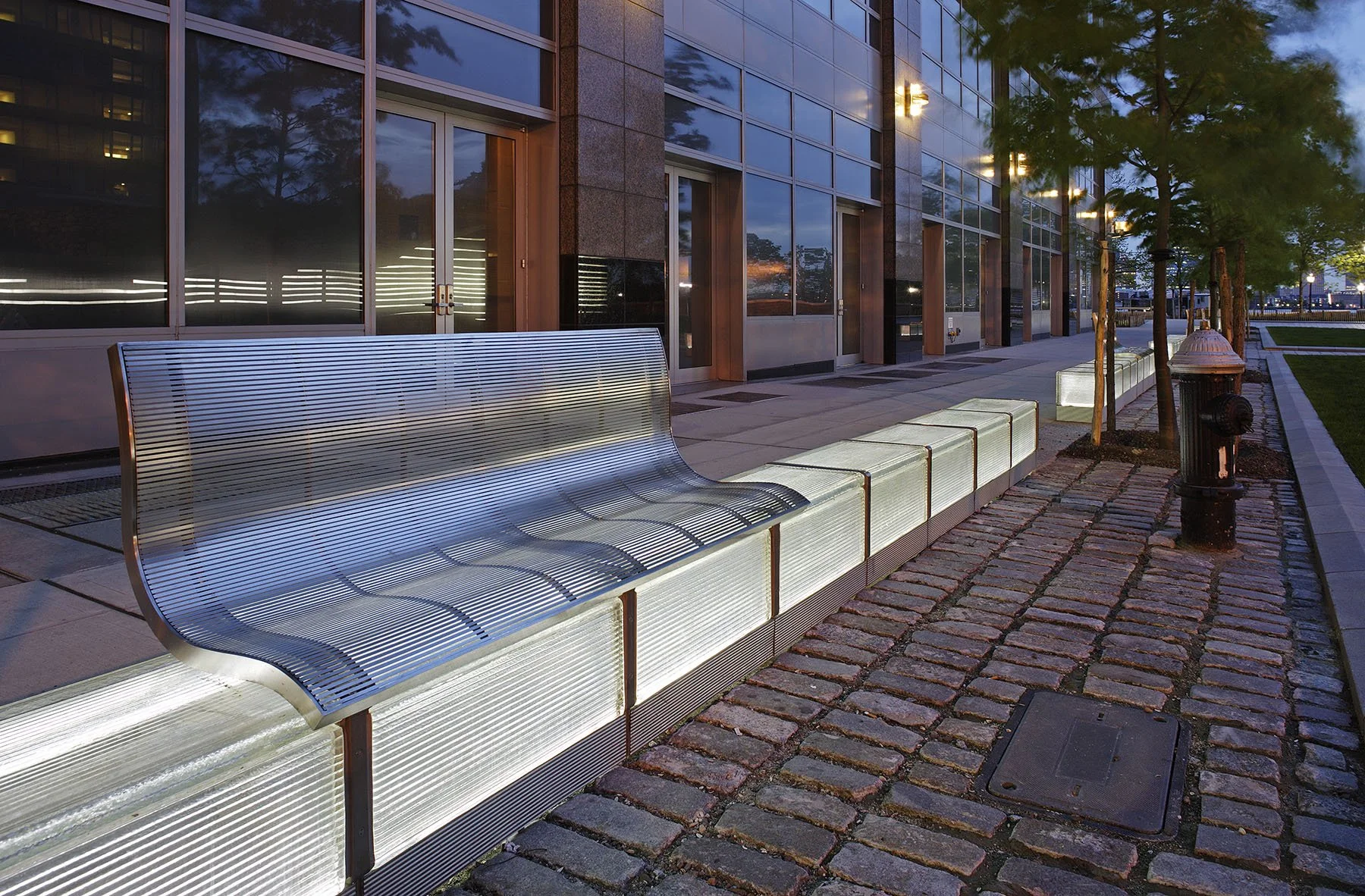 Close-up of a curved metal bench with wave-patterned design on a cobblestone streetscape in Battery Park City.
