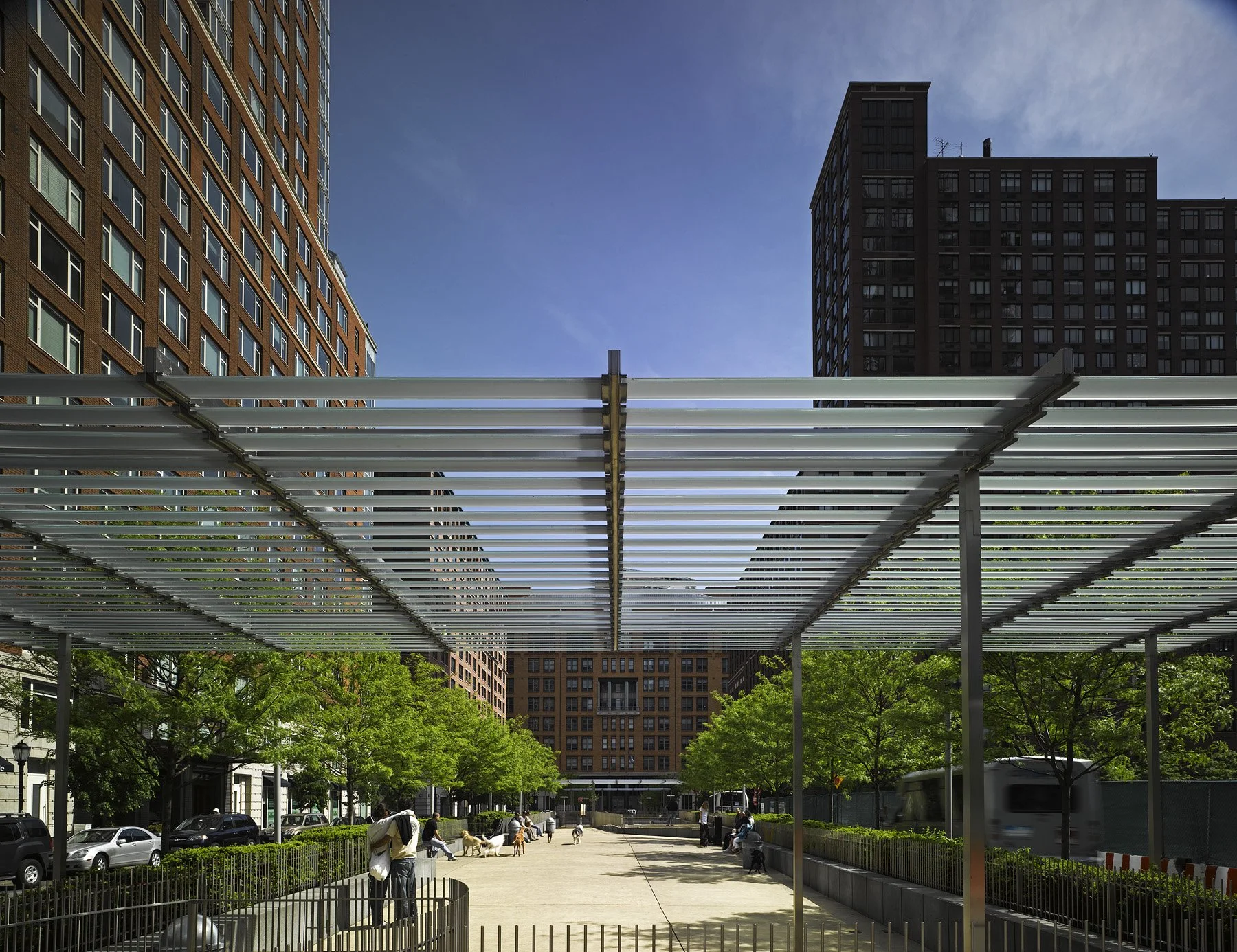 Daytime view of a shaded pergola walkway with trees and seating in Battery Park City streetscape.