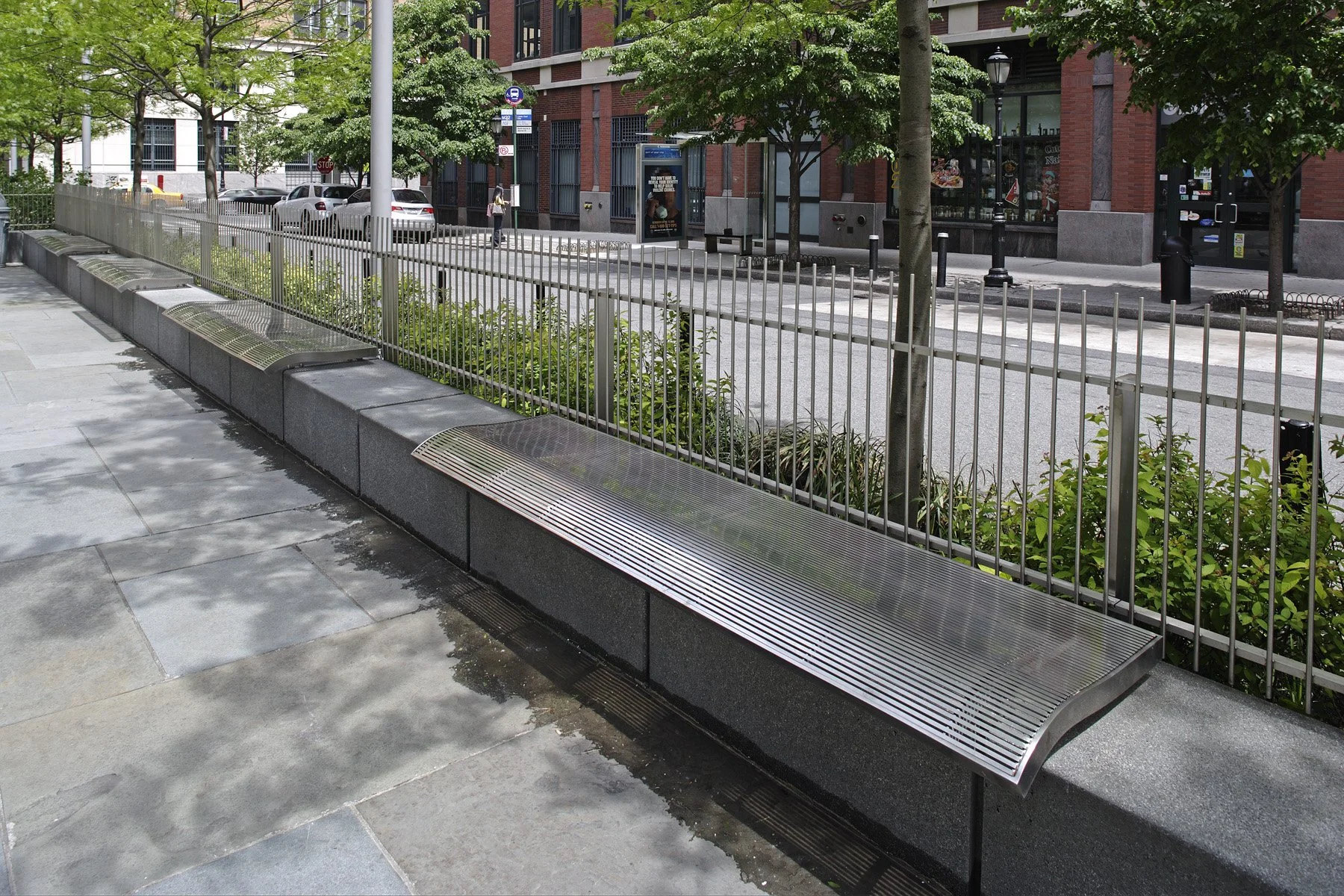 Curved metal bench alongside a planted green border on a Battery Park City pavement.