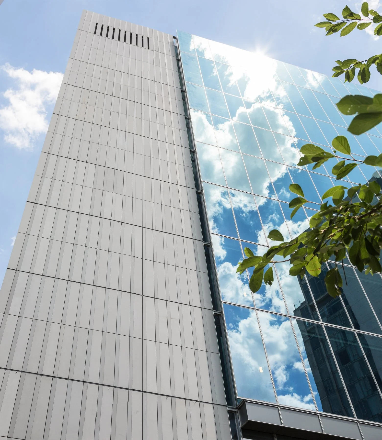Looking up at a modern building facade with stone cladding and glass curtain wall reflecting clouds and blue sky.