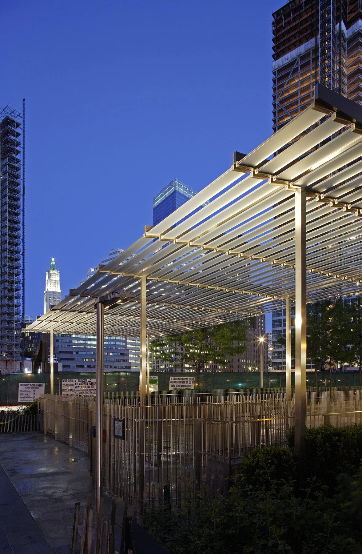 Metal pergola canopy structure lit at dusk with a church steeple visible in the Battery Park City skyline.