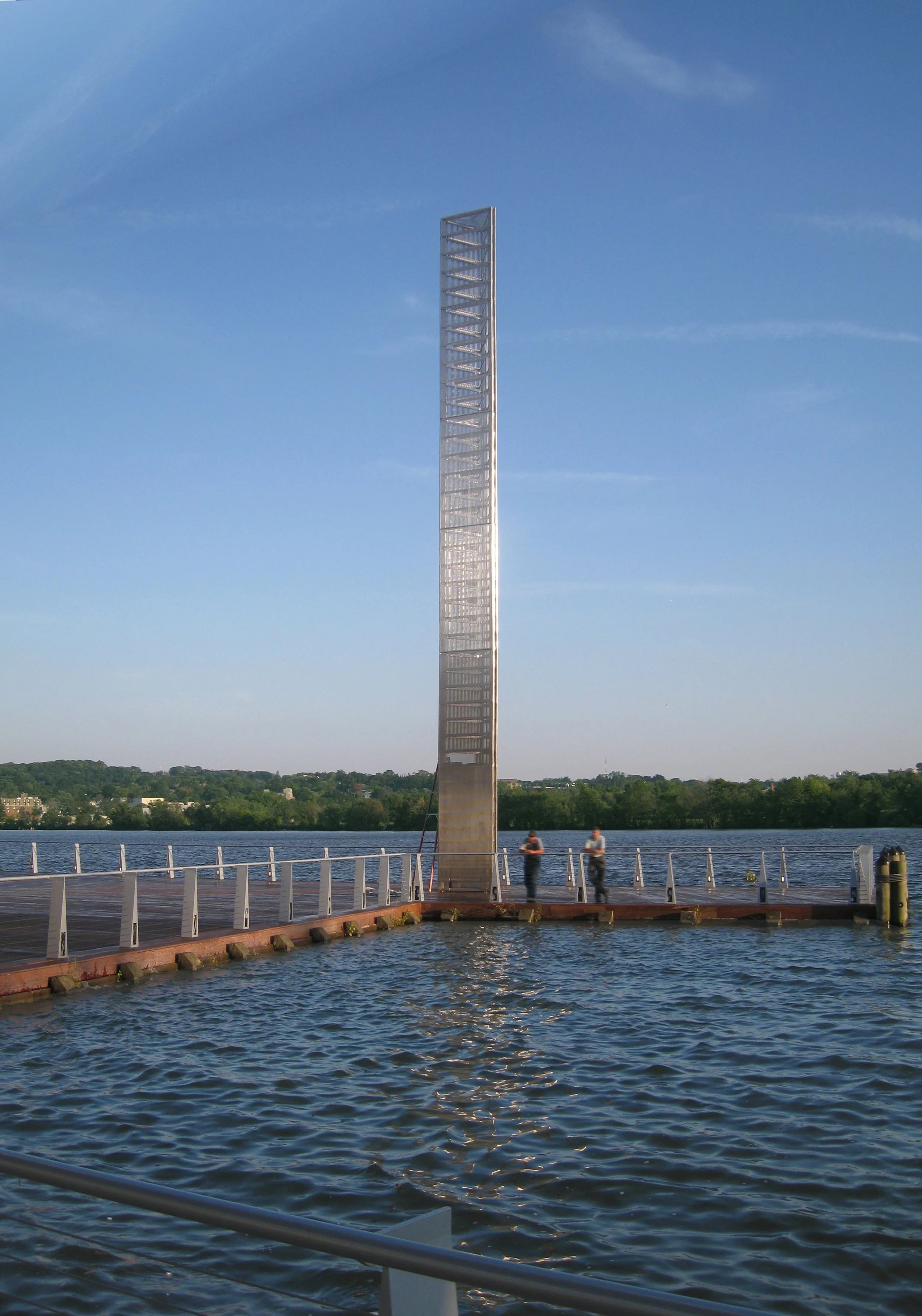 Prismatic Light Pillar rising above a calm waterfront with clear blue sky in the background.