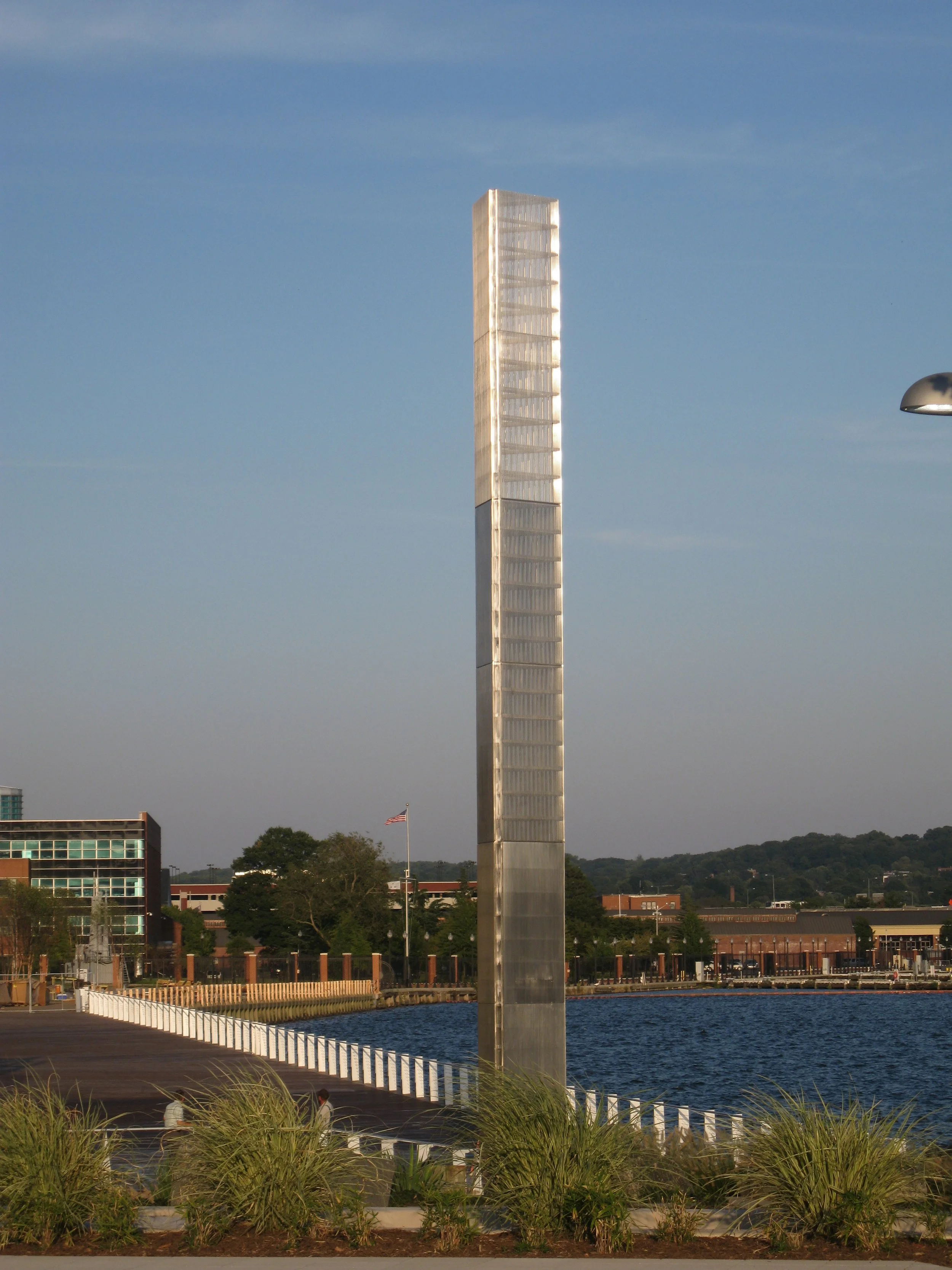 Tall reflective Prismatic Light Pillar sculpture standing beside a waterfront on a clear day.