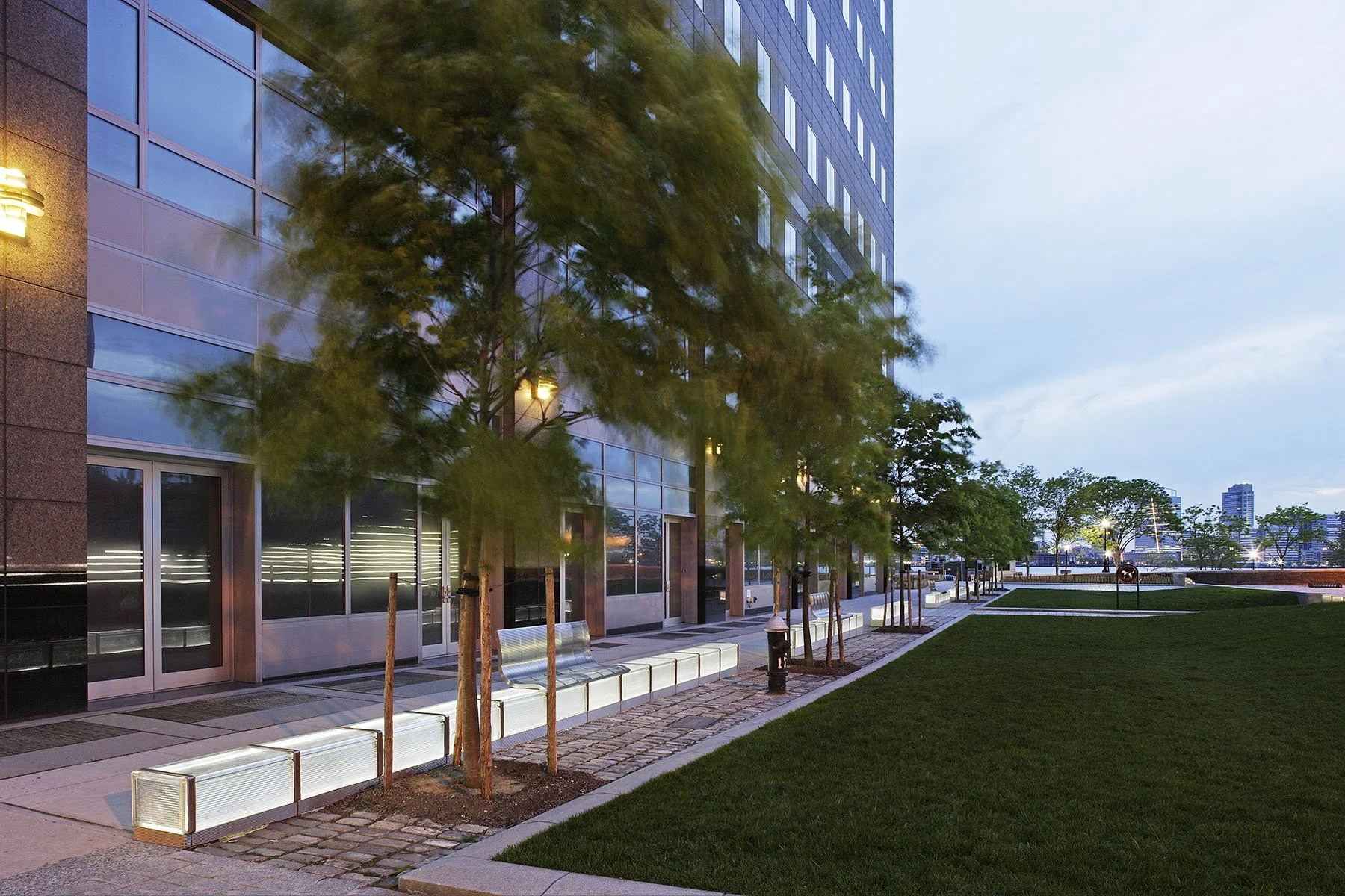 Tree-lined pedestrian streetscape at dusk with modern street furniture along a Battery Park City boulevard.