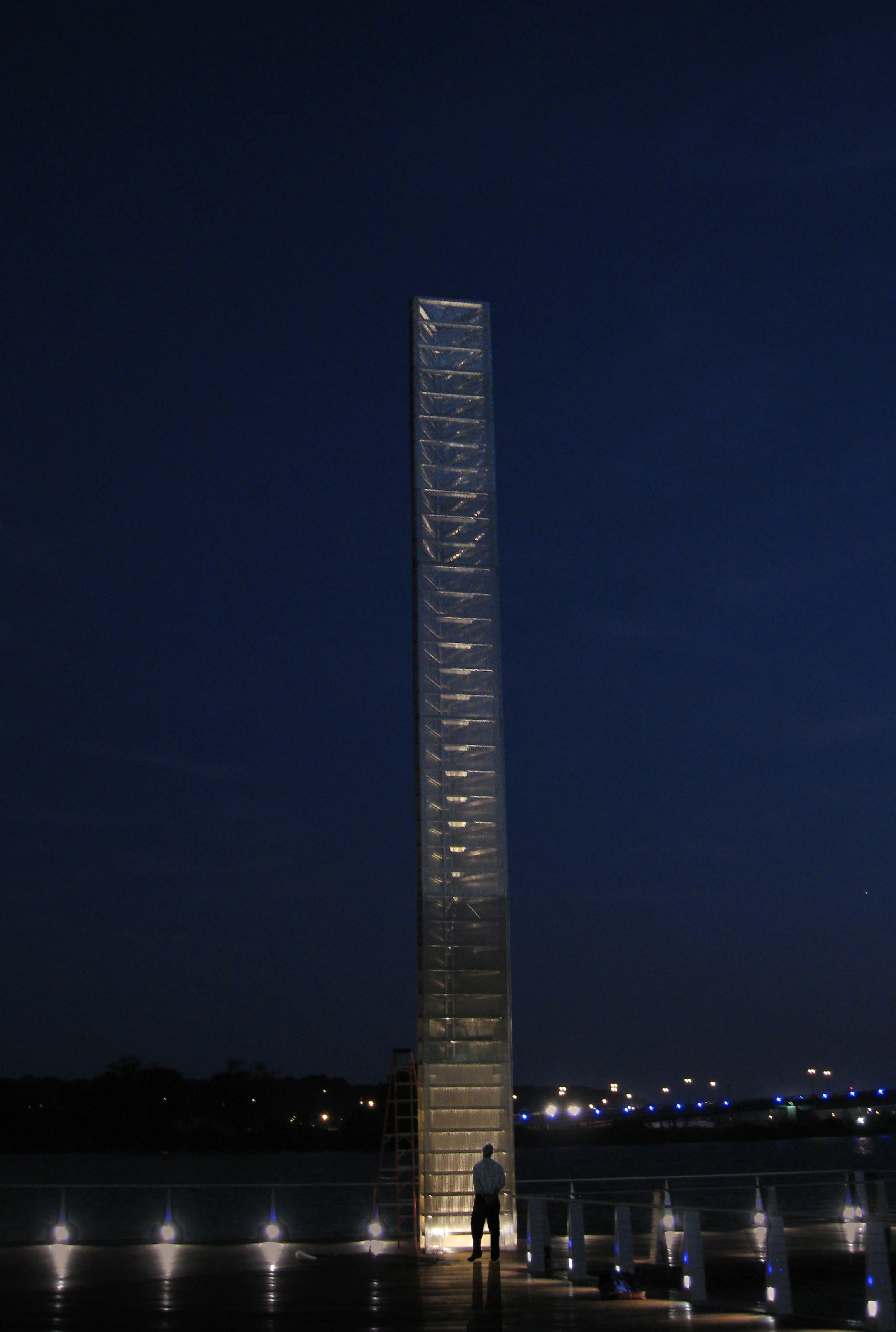 Prismatic Light Pillar sculpture illuminated against a dark evening sky at a waterfront location.