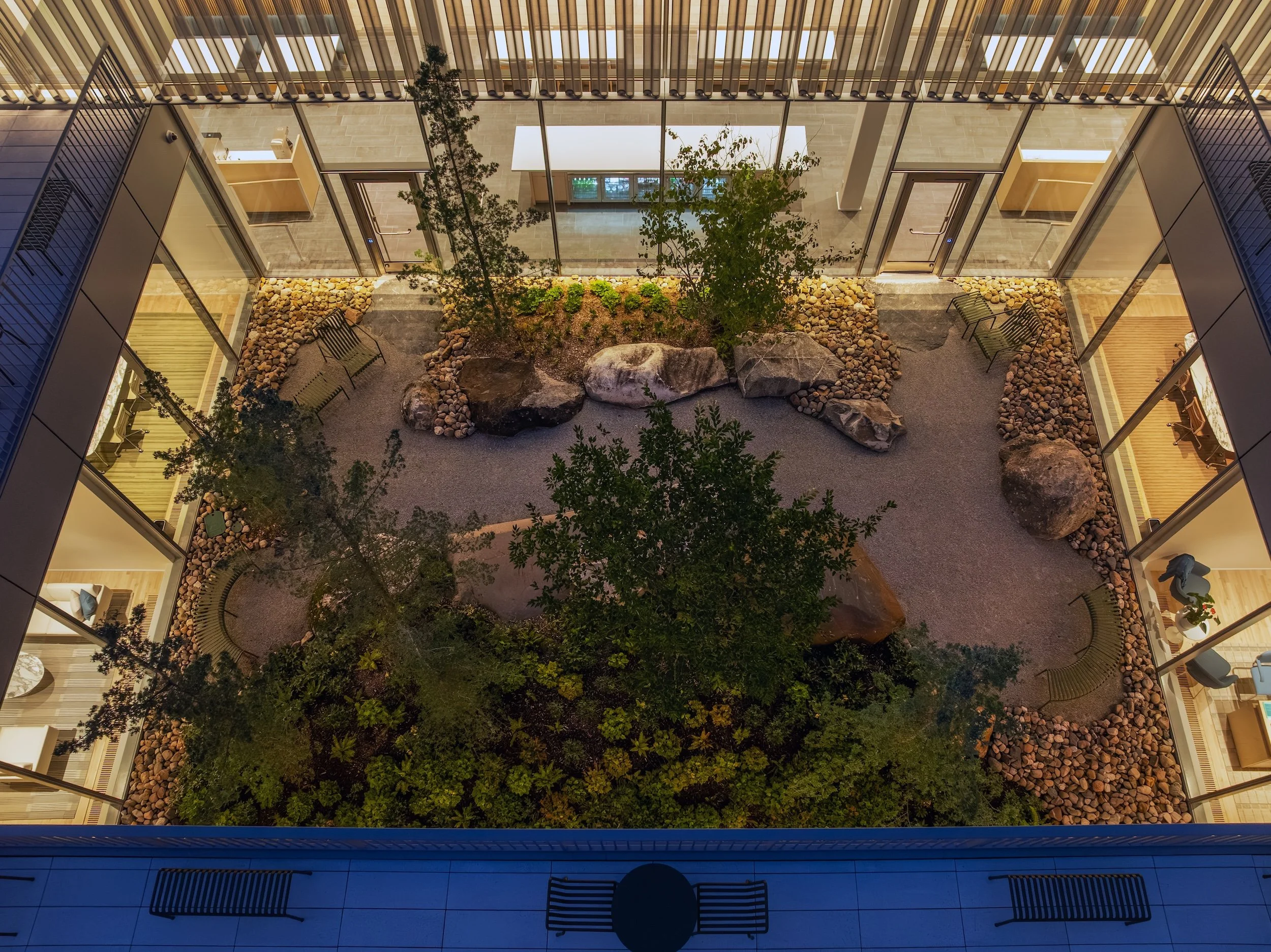 Overhead view of a modern indoor courtyard featuring landscaped greenery, trees, rocks, and gravel pathways surrounded by glass-walled rooms.