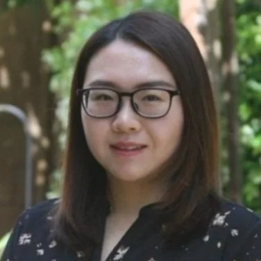 A woman with glasses and shoulder-length dark hair smiling outdoors among trees.