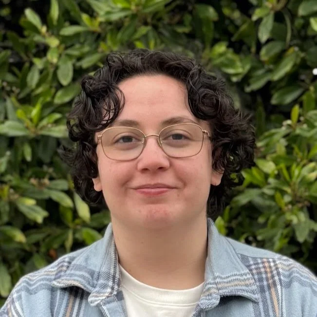Young person with short curly dark hair and glasses, wearing a light plaid shirt, standing outdoors in front of green foliage.