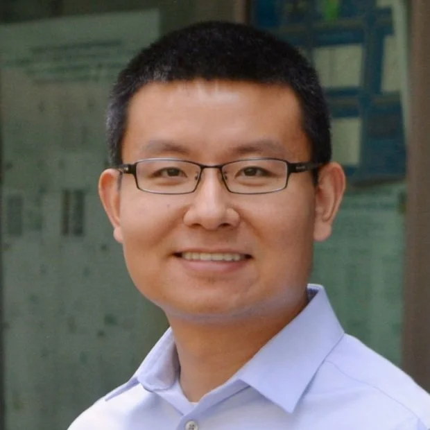 A smiling man with short black hair, glasses, and a light blue collared shirt, standing outdoors with a blurred building in the background.