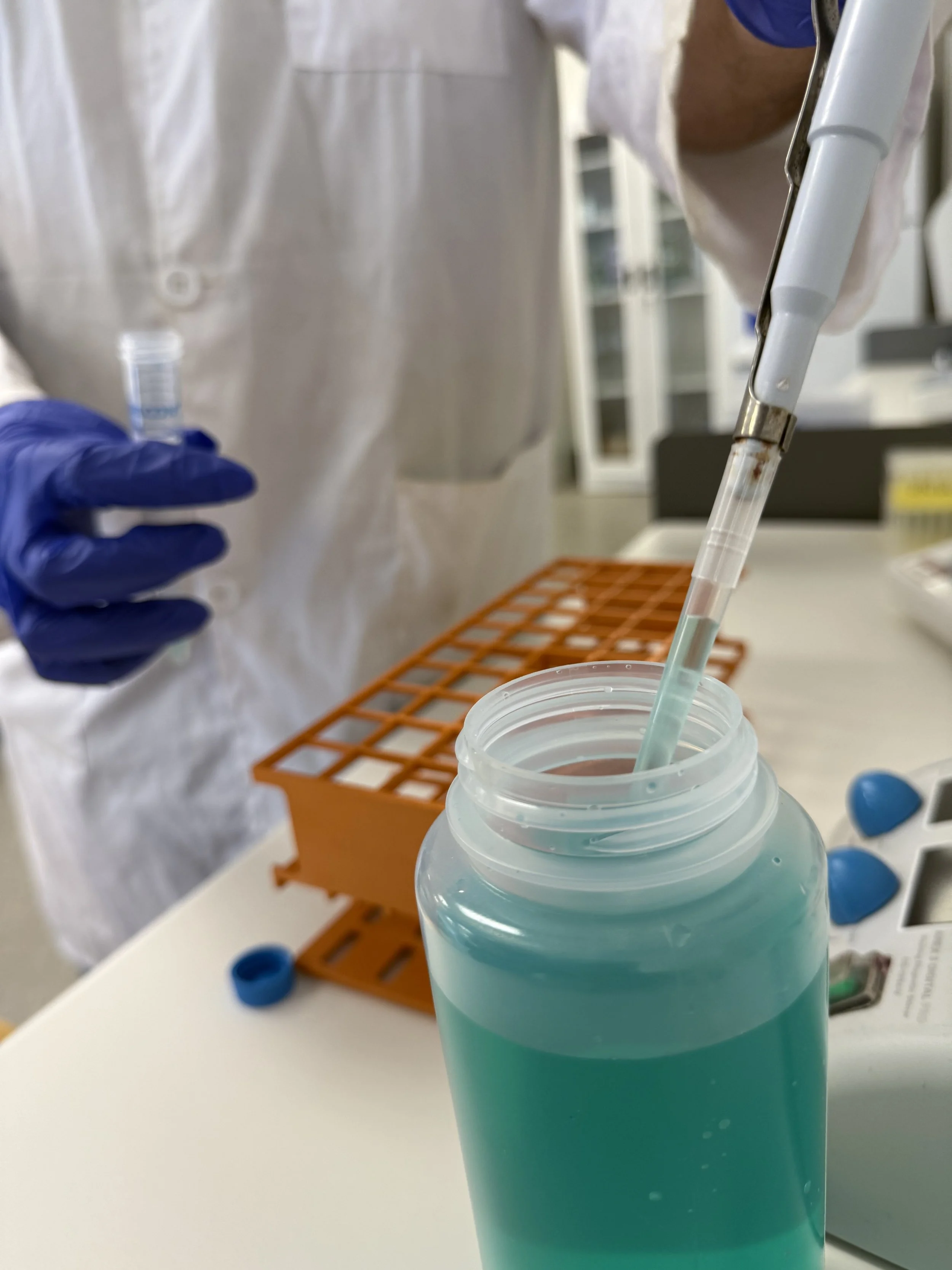 A scientist in a white lab coat and blue gloves dispensing a blue liquid into a test tube in a laboratory setting.