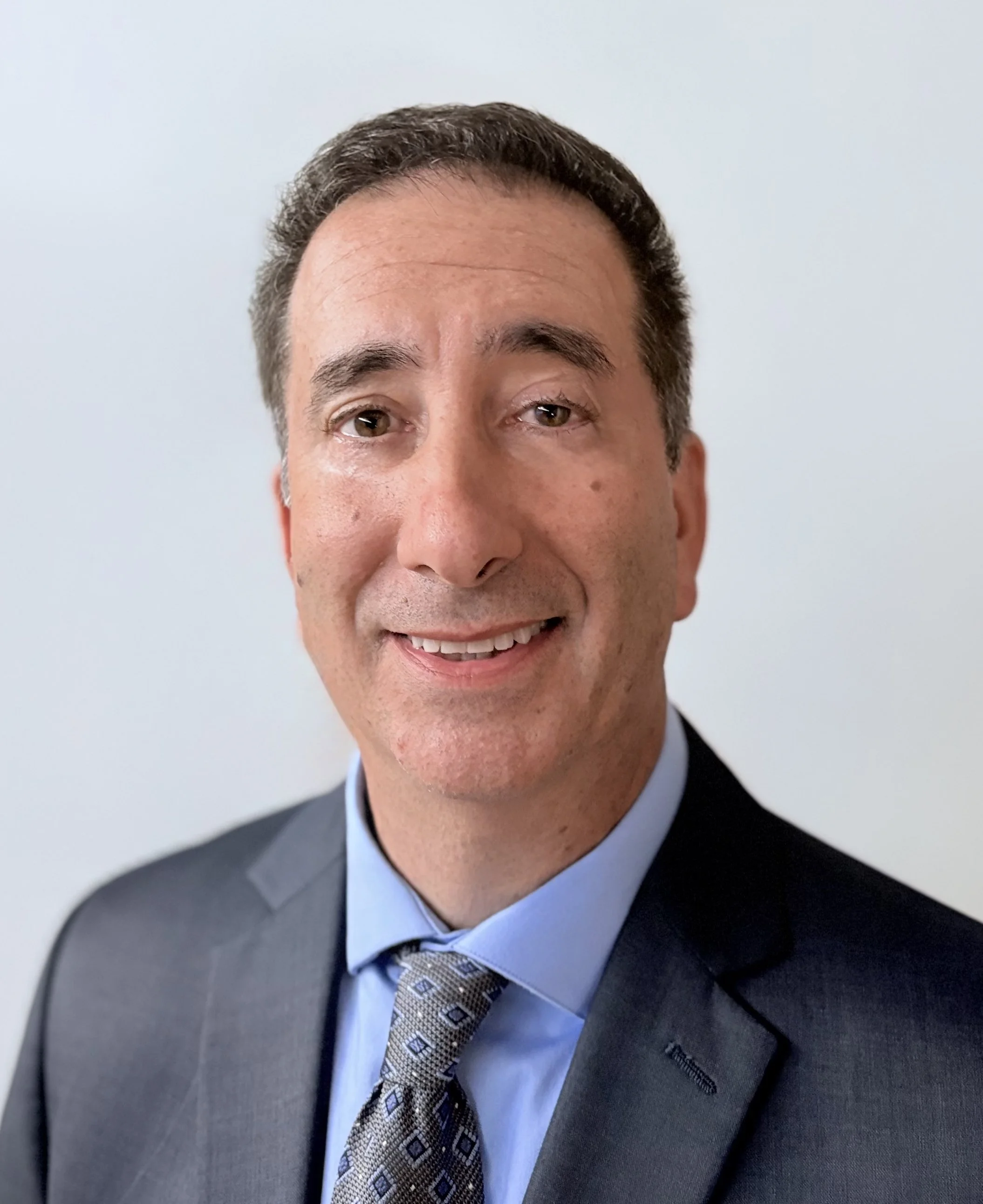 A professional headshot of a man with dark hair, wearing a dark suit, light blue shirt, and patterned tie, smiling against a plain light-colored background.