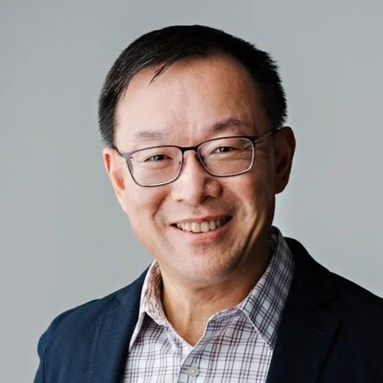 A smiling man with glasses wearing a checked shirt and a dark blazer