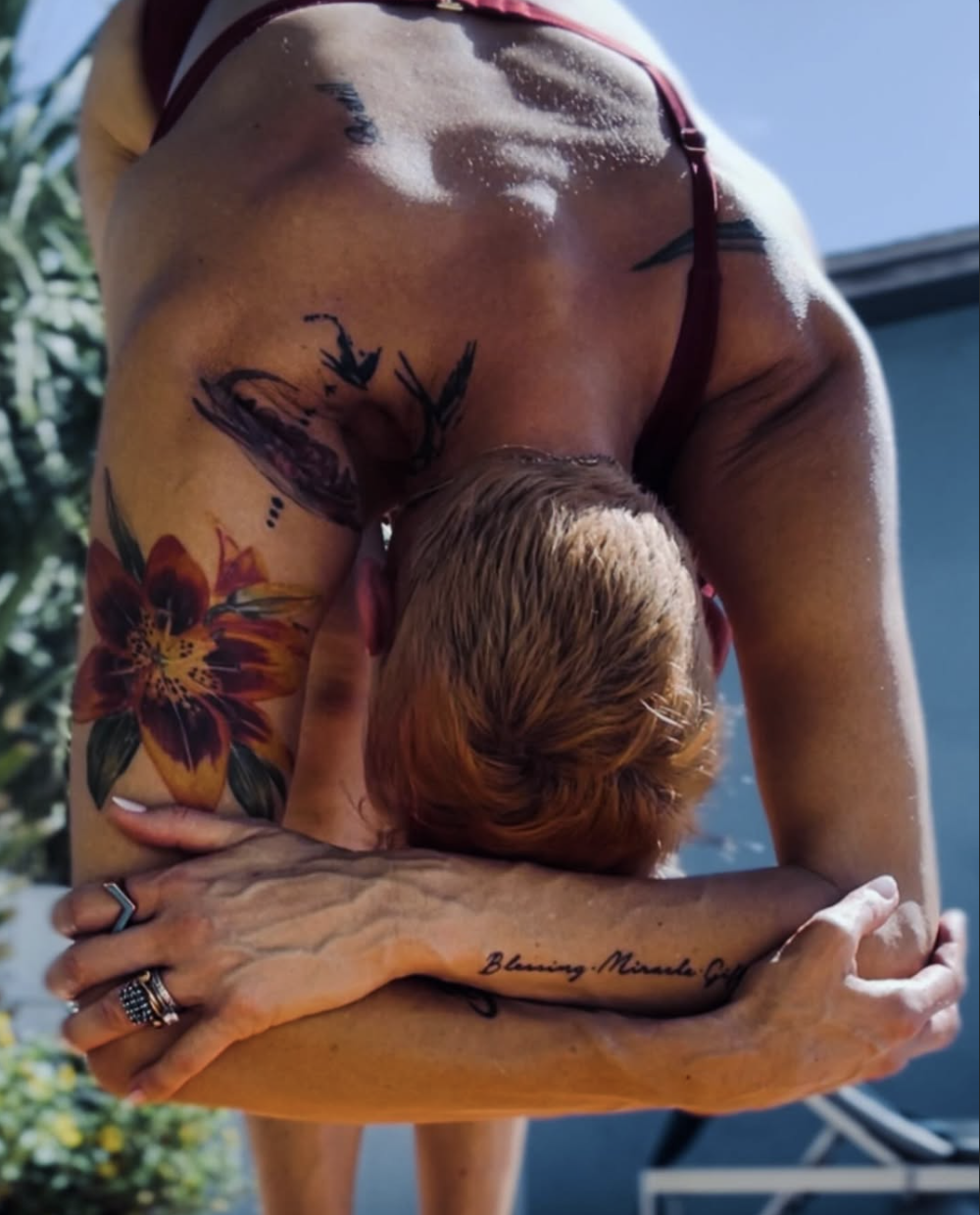 Person with tattoos doing a forward bend yoga pose outdoors, showing back and arm tattoos, wearing a maroon top, with sunlight and greenery in the background.
