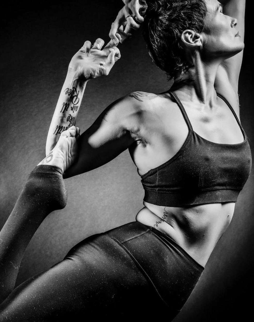 Black and white photo of a woman in athletic wear stretching her arm behind her head, with another person's hands assisting her with her stretch. The woman has tattoos on her arm and side, and short dark hair.