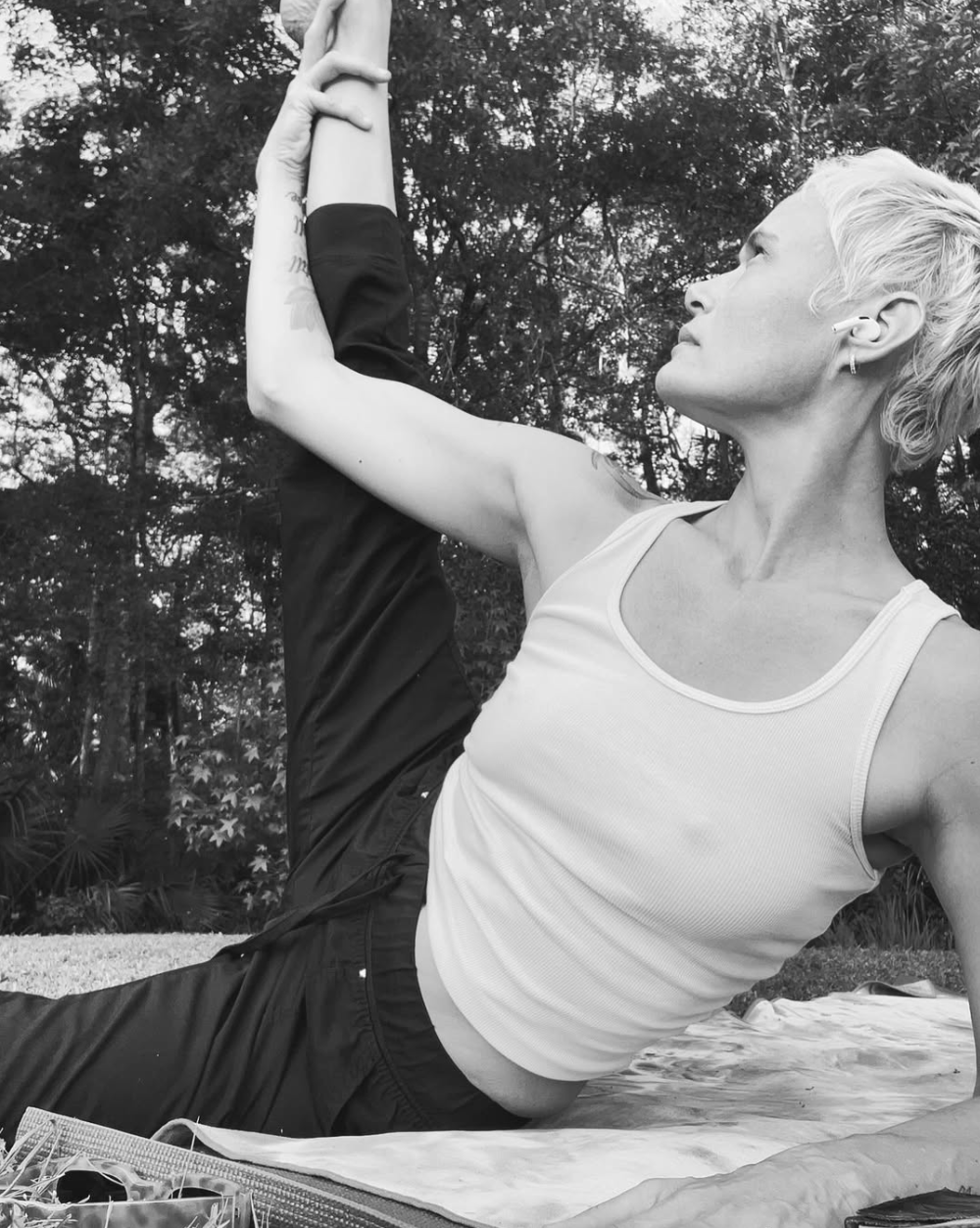 A woman doing yoga outdoors, lying on a mat with trees in the background, stretching her arm upward and to the side.