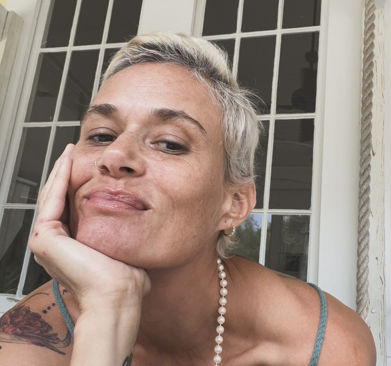 A woman with short, platinum blonde hair and a nose ring resting her chin on her hand. She has a tattoo on her arm and is wearing a pearl necklace and a teal top. She is sitting outside near a window with a white frame.