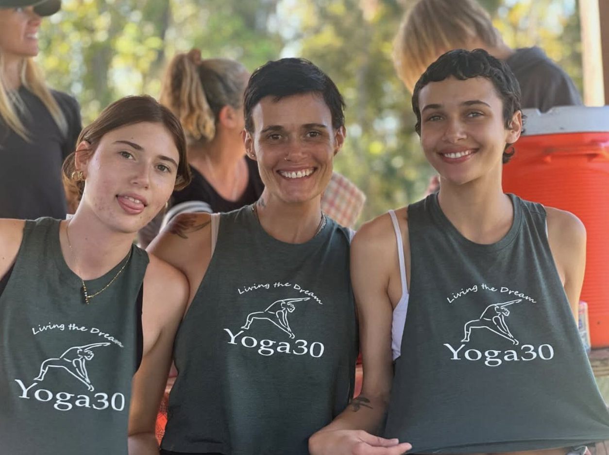 Three women in yoga tank tops smiling outdoors, with two in the background, at a yoga event or gathering.